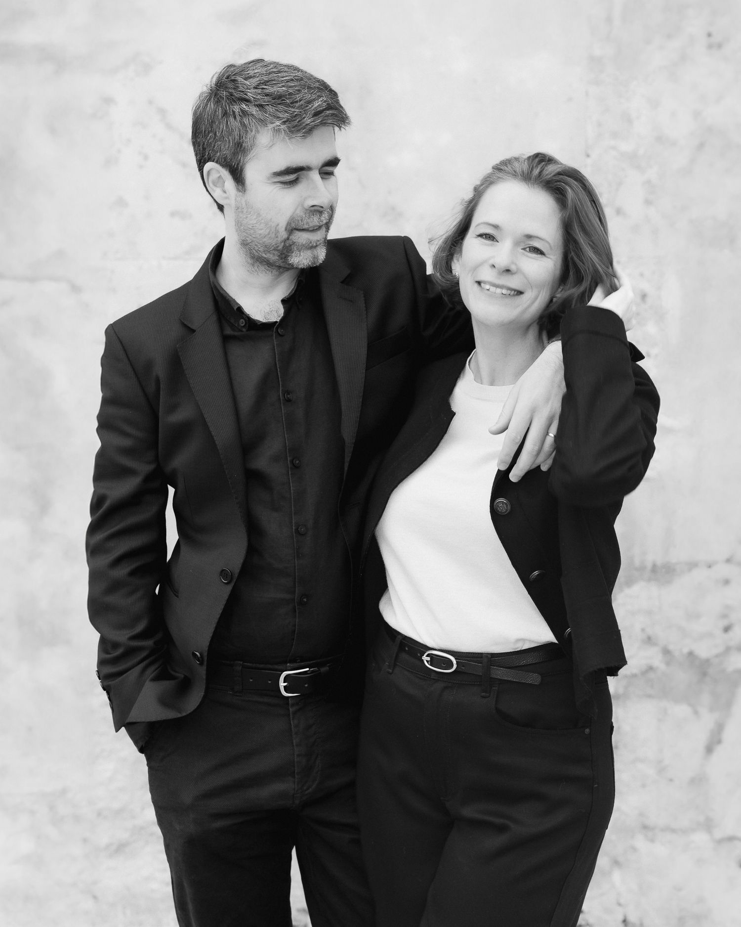 Black and white portrait of a couple in dark business attire smiling together against a light wall.