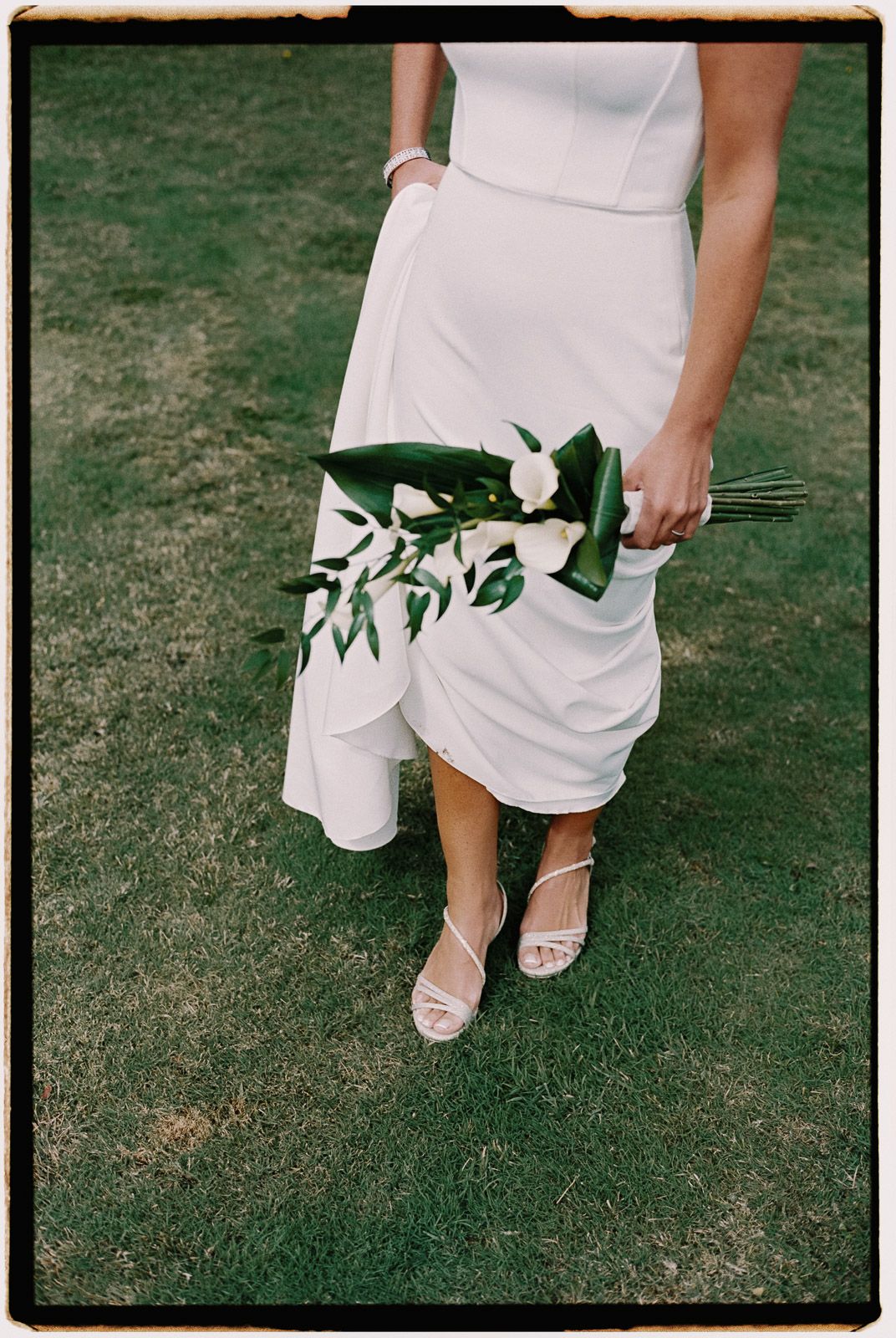 Bride in white dress holding minimalist bouquet of white flowers and green leaves, standing on green grass.