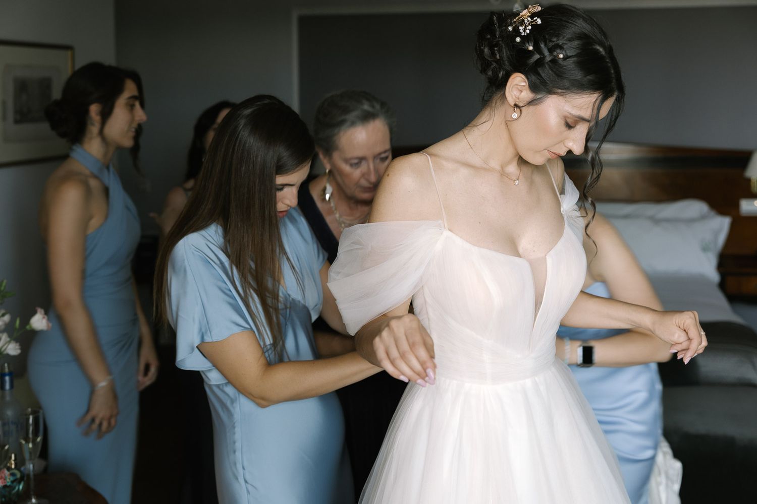 Bridesmaids in blue dresses helping bride with her off-shoulder white tulle wedding gown in a hotel room.