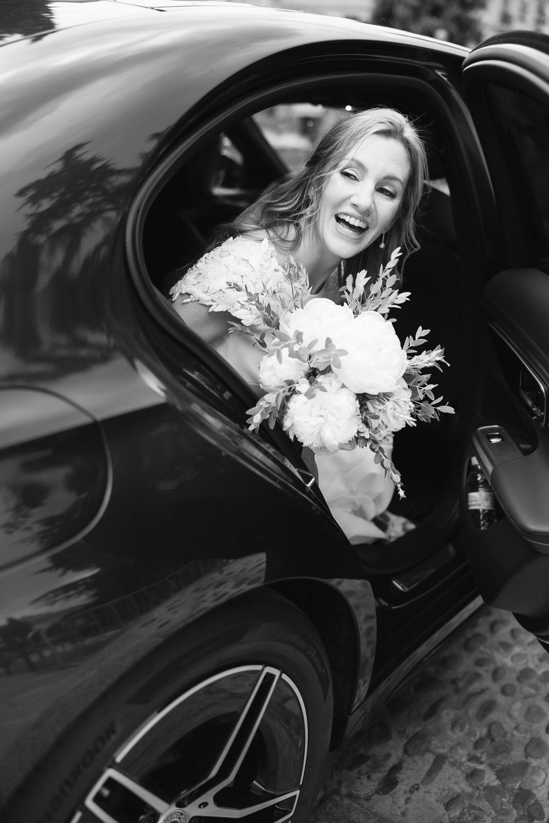 Joyful bride holding a white bouquet laughing from a luxury car on her wedding day.