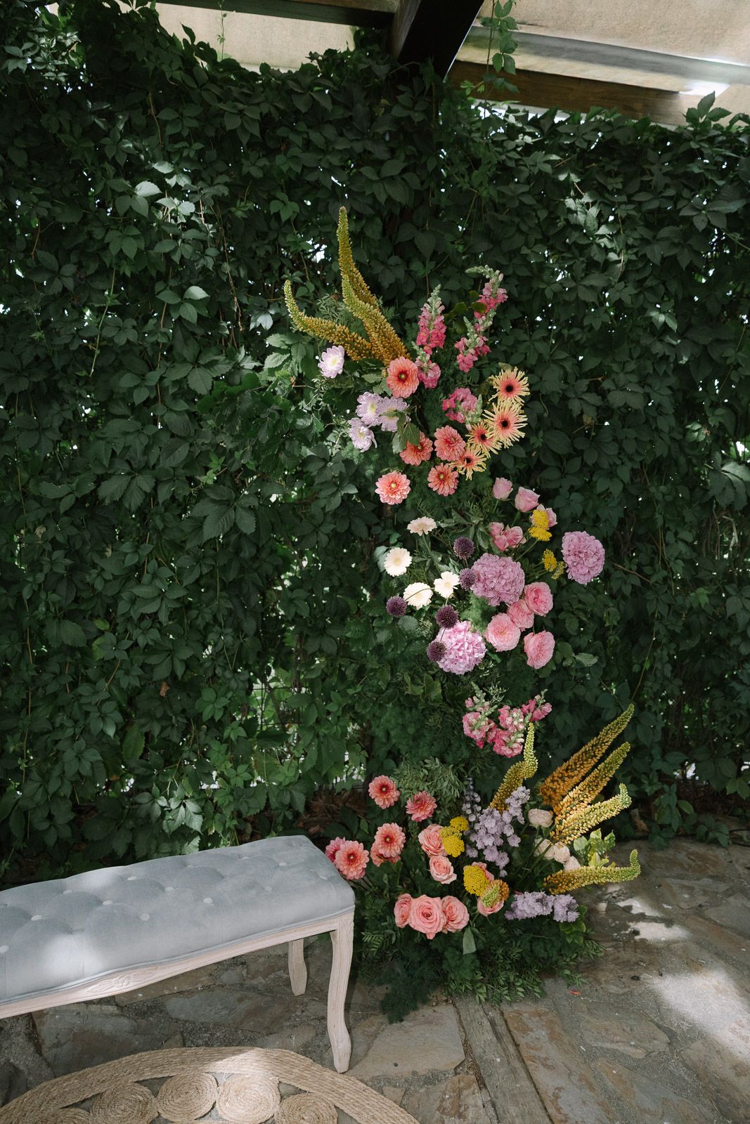 Lush floral wedding installation with pink, orange, and gold blooms against a green ivy wall beside a white bench.