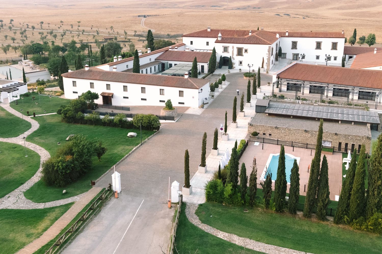 Aerial view of Mediterranean villa resort with white buildings, cypress trees, swimming pool and winding paths in rural setting.