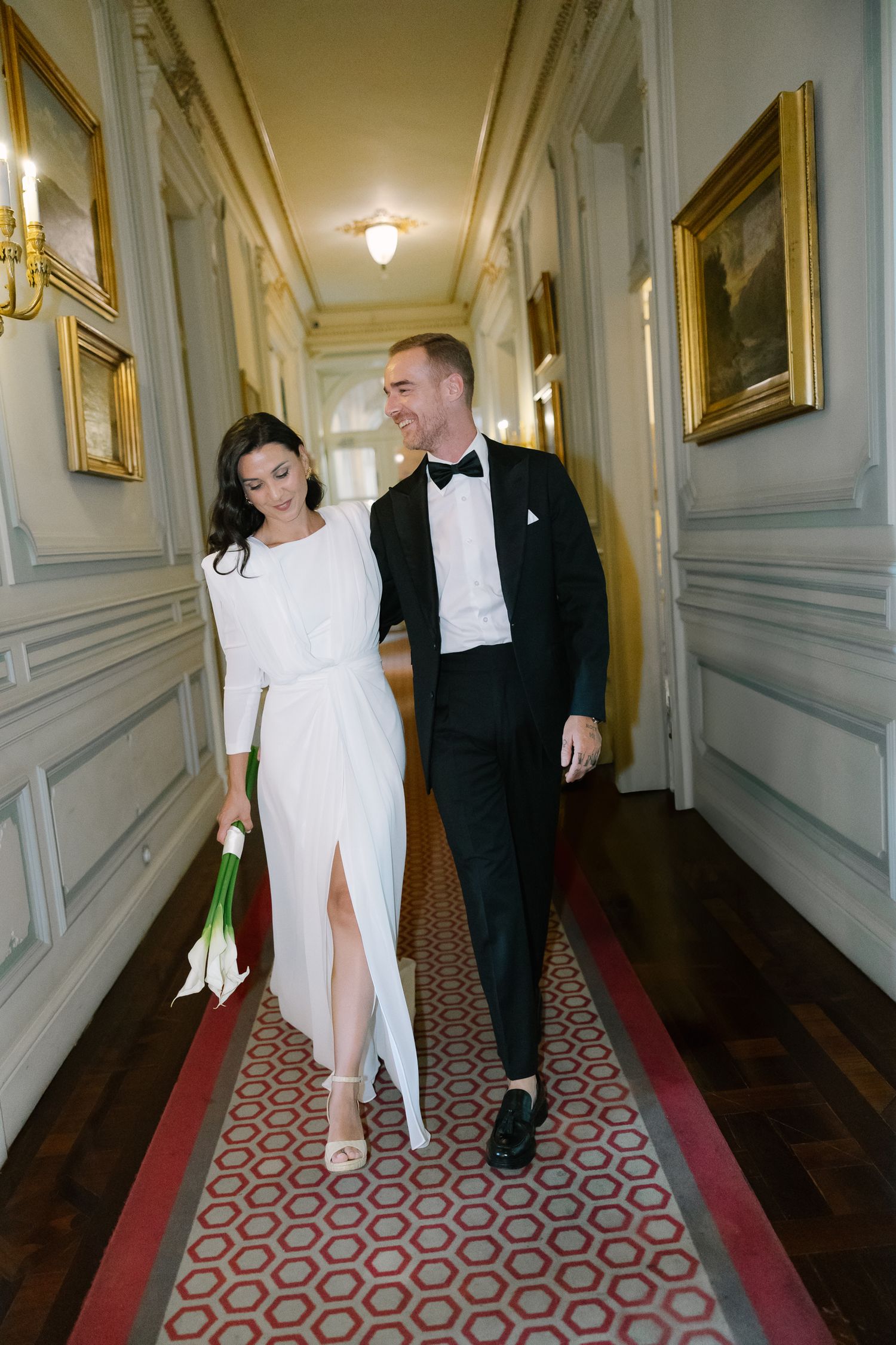 A couple in formal attire walking down an elegant hallway with patterned carpet and gold-framed artwork on walls.