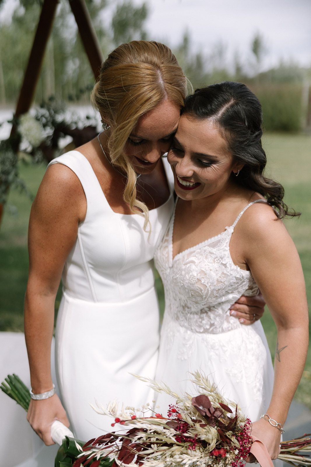 Two brides in white wedding dresses embrace and smile while holding a bouquet of burgundy and dried flowers outdoors.