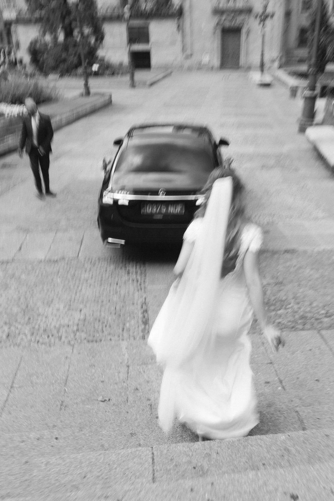 Black and white photo of a bride in white dress walking toward a black car on cobblestone street, groom visible in background.