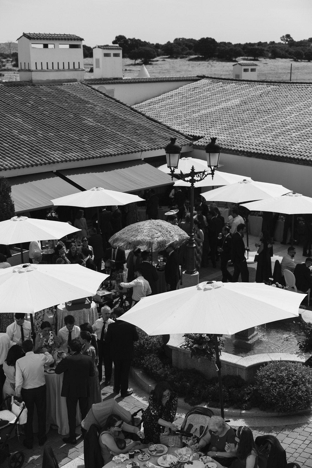 Black and white overhead view of elegant outdoor wedding cocktail hour with white market umbrellas on terracotta roof terrace.