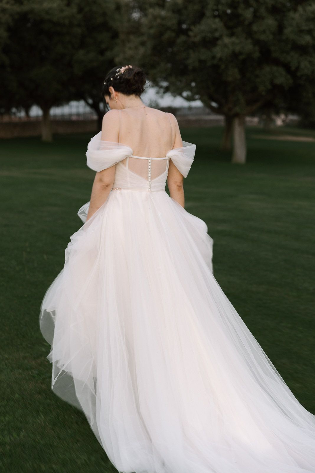 Bride in elegant off-shoulder white tulle ballgown with button back detail, standing on green lawn near trees.