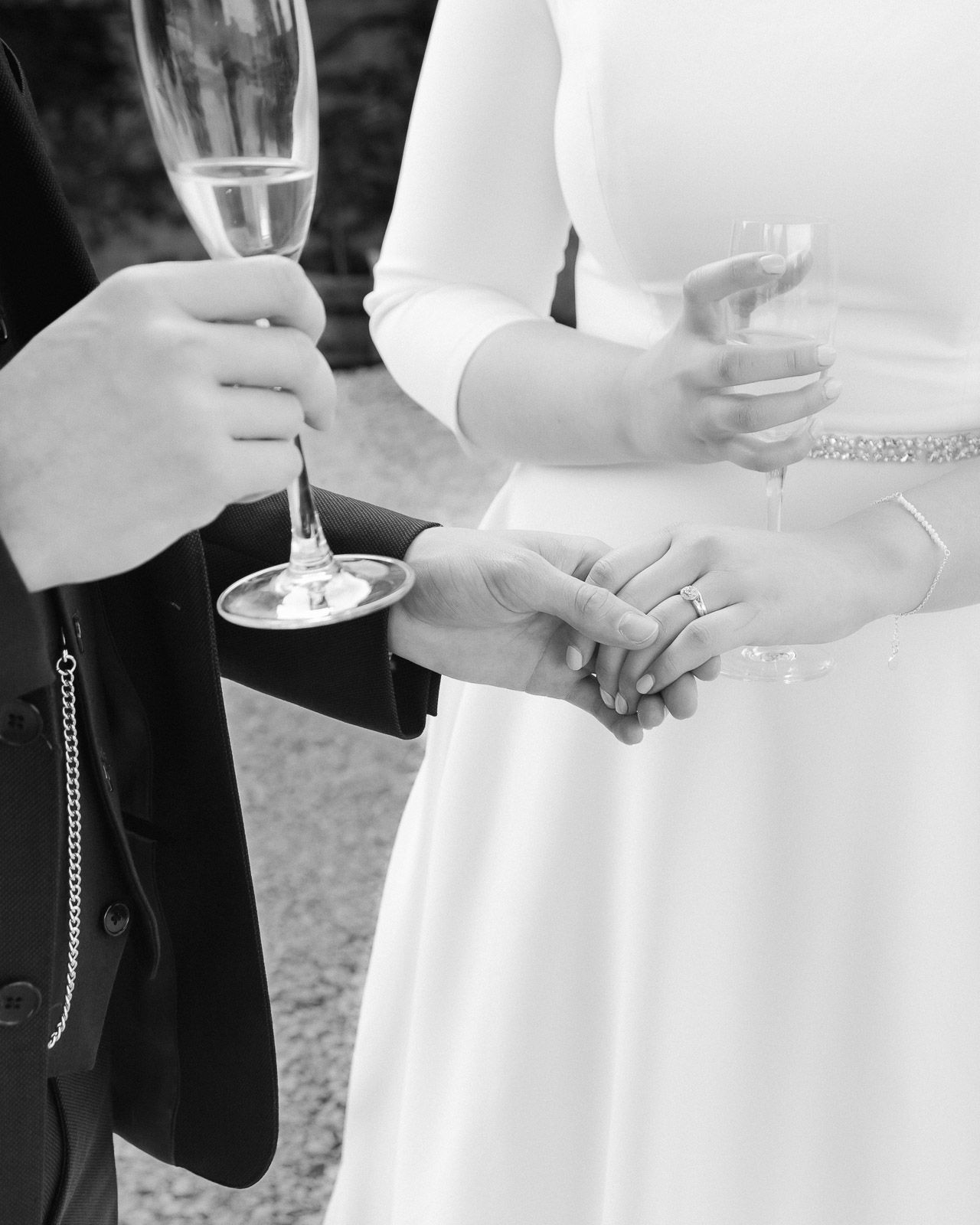 Black and white photo of newlyweds holding hands and champagne glasses, bride in white dress with wedding ring visible.