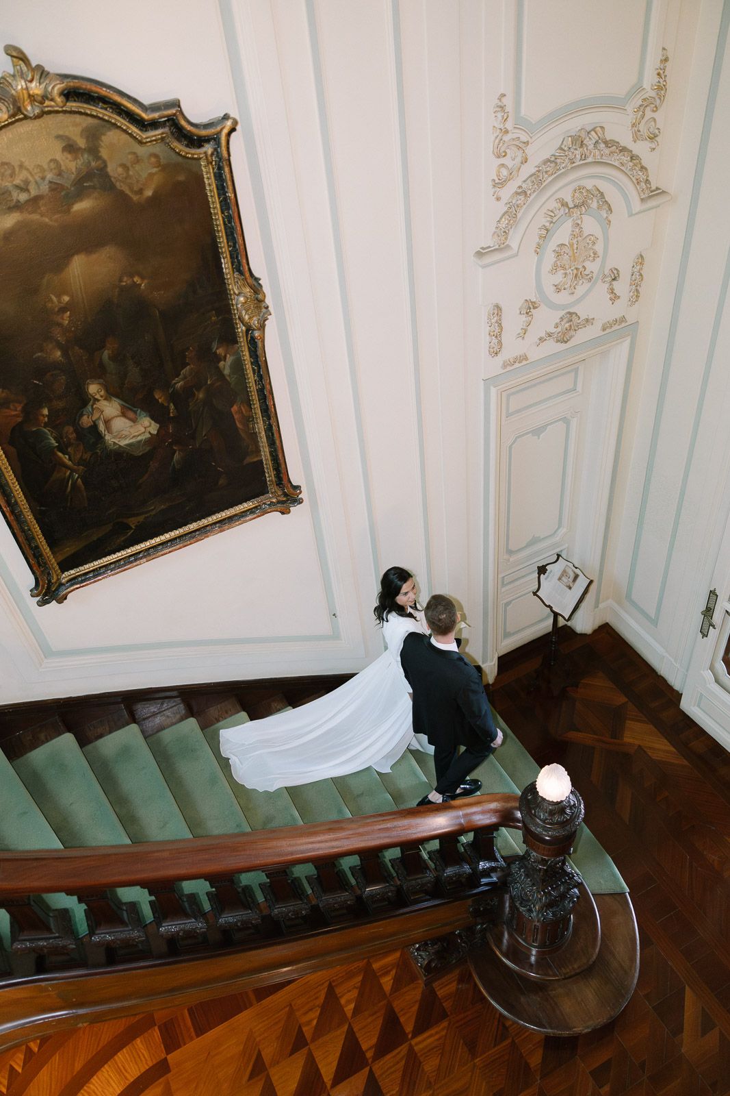 Bride and groom walking up elegant staircase in ornate white ballroom with classical painting and green carpet.