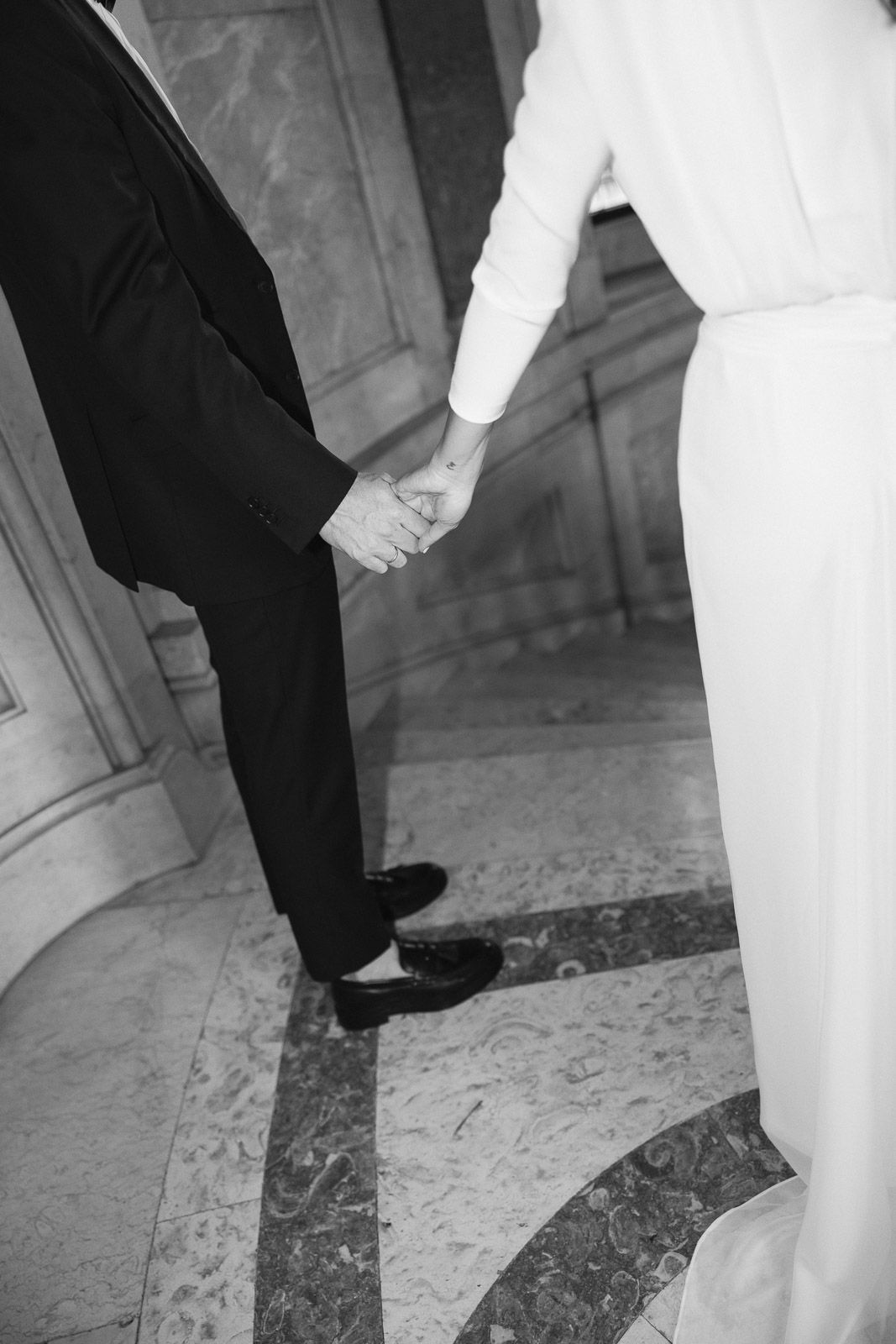Black and white photo of couple holding hands in formal wear on marble floor at wedding.