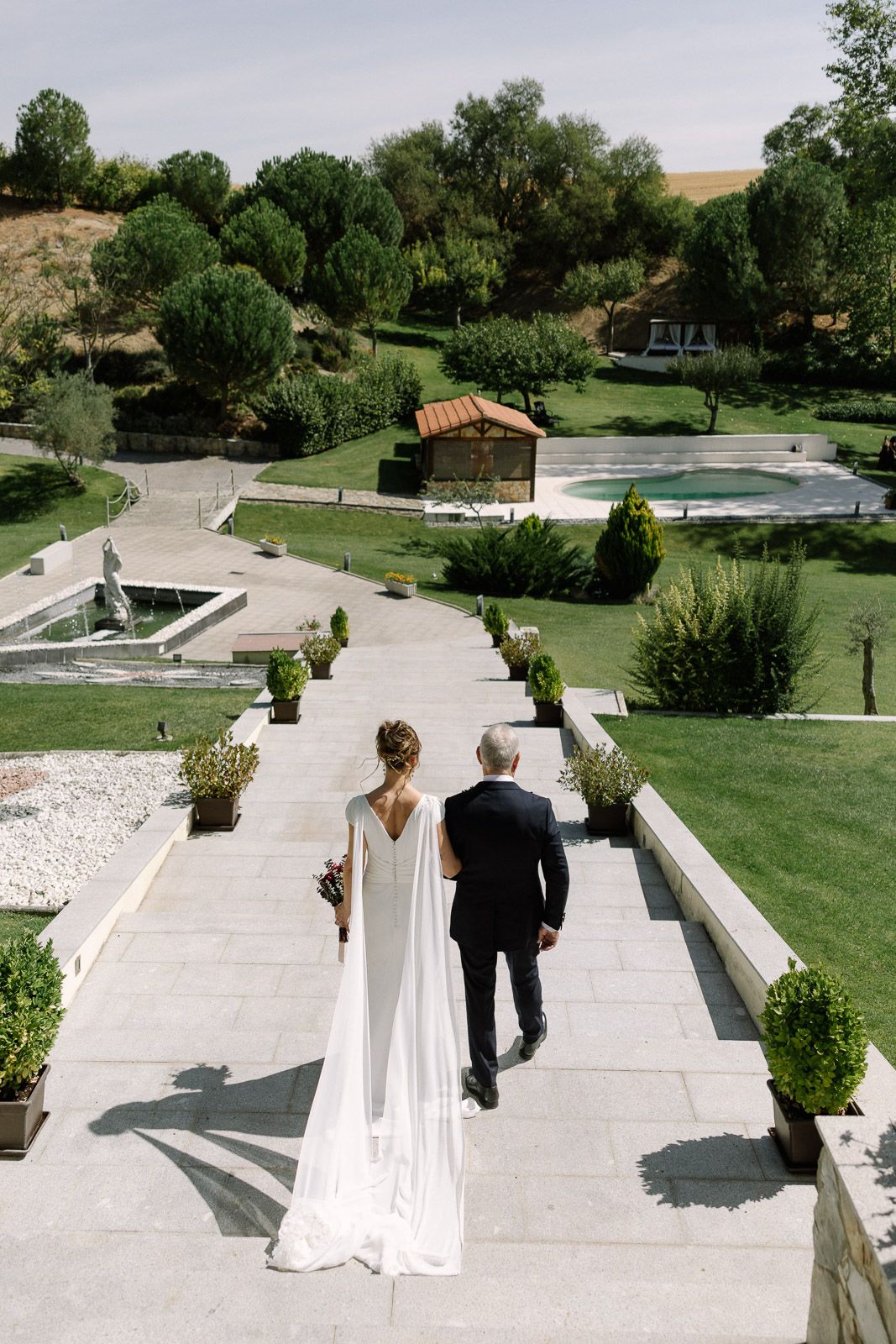 Bride in elegant white gown walks with groom through manicured garden estate on their wedding day.