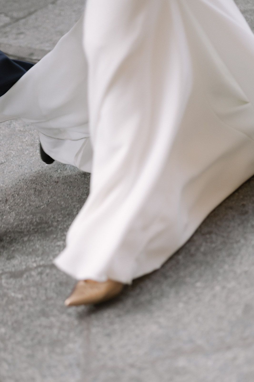 A bride in a flowing white wedding gown and nude heels walks gracefully on a gray pavement.