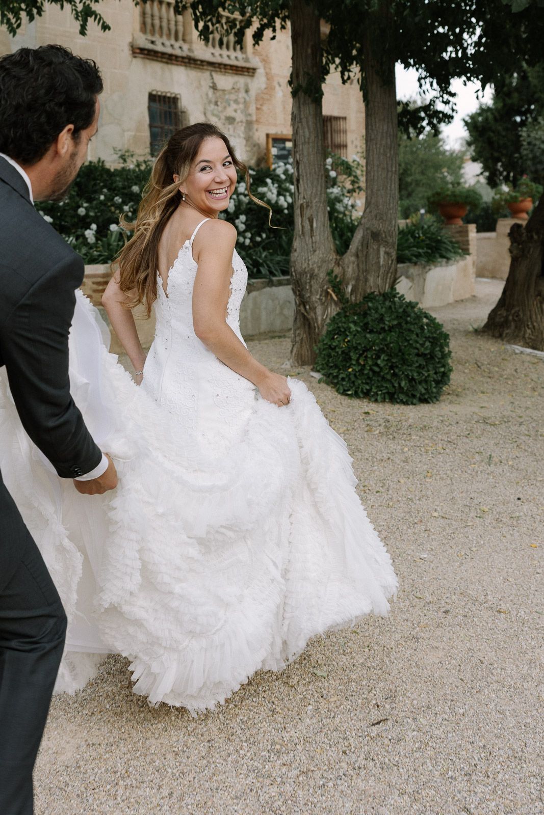 Bride in a white ruffled gown smiles back at groom in a suit while walking through a garden venue.