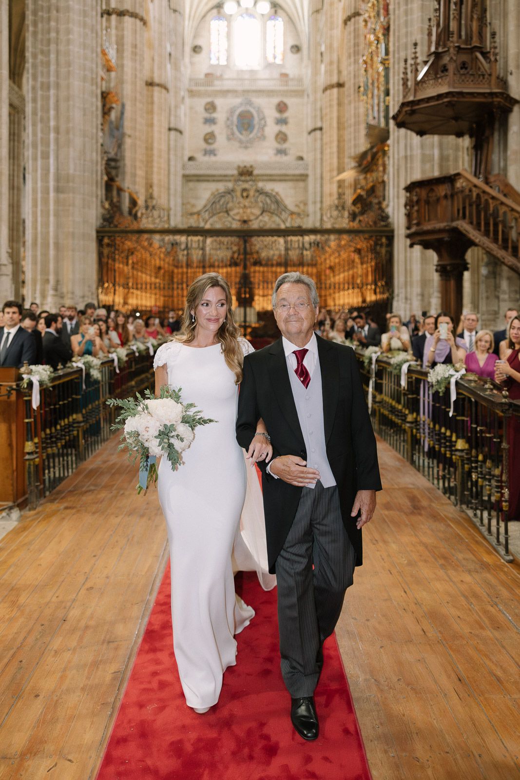 Bride in white gown walks down cathedral aisle with escort on red carpet, guests seated in ornate church.