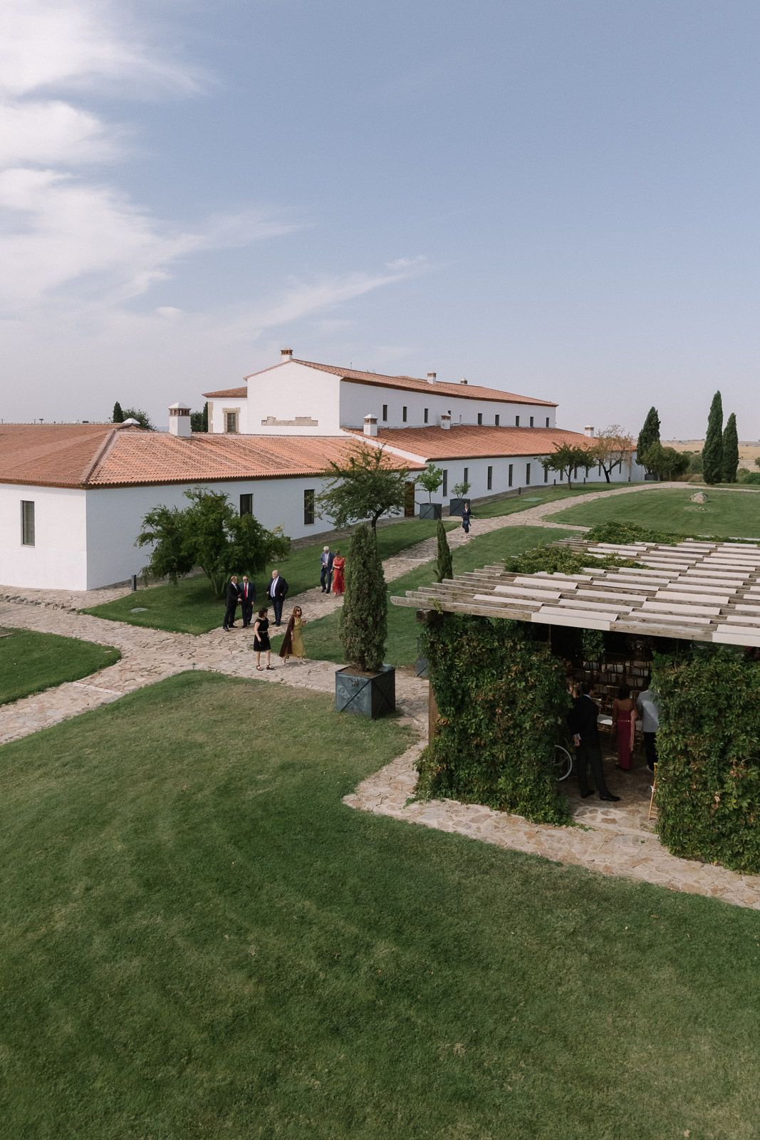 Aerial view of elegant white Spanish wedding venue with terracotta roof, manicured lawns, and cypress trees under blue sky.