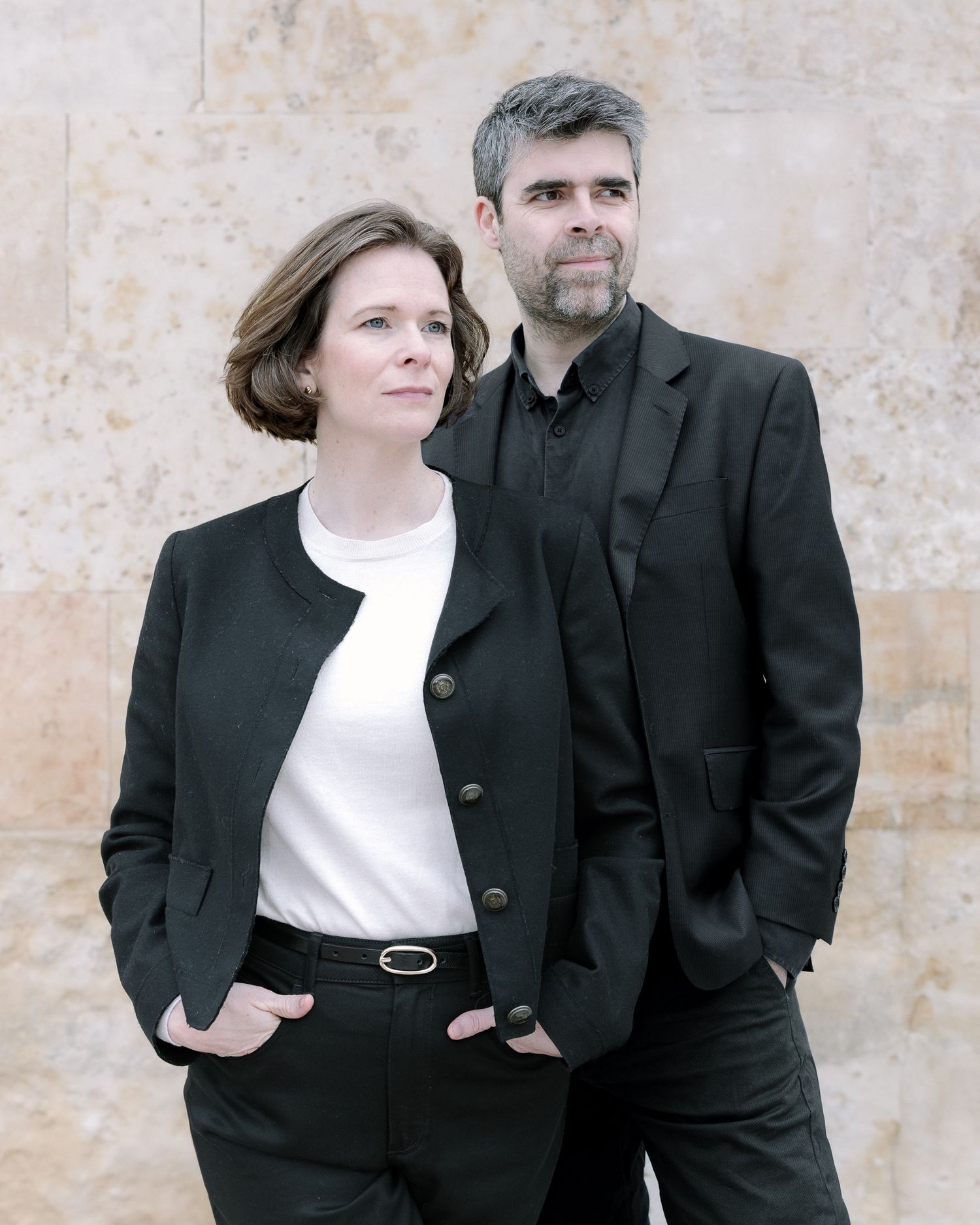 Two people in matching black blazers and formal attire pose together against a textured wall.