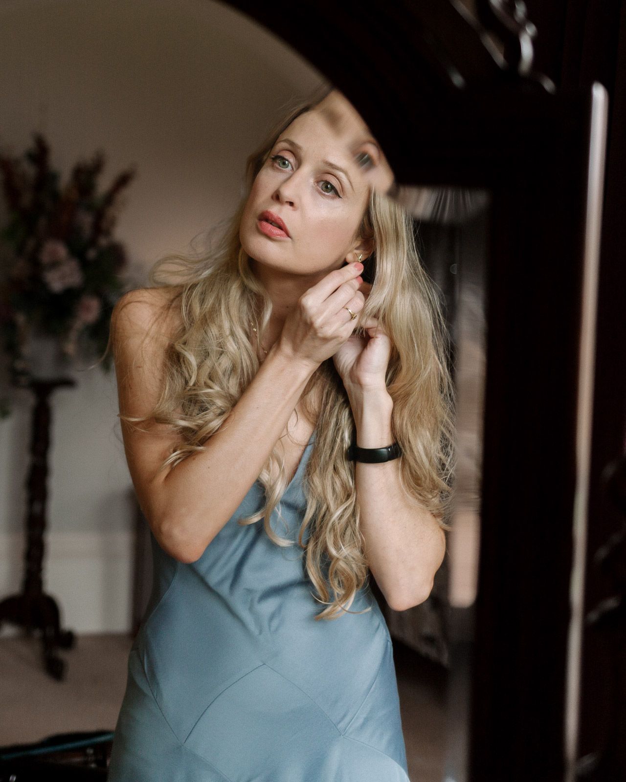 Elegant blonde woman in blue slip dress adjusting earring by mirror in dimly lit room.