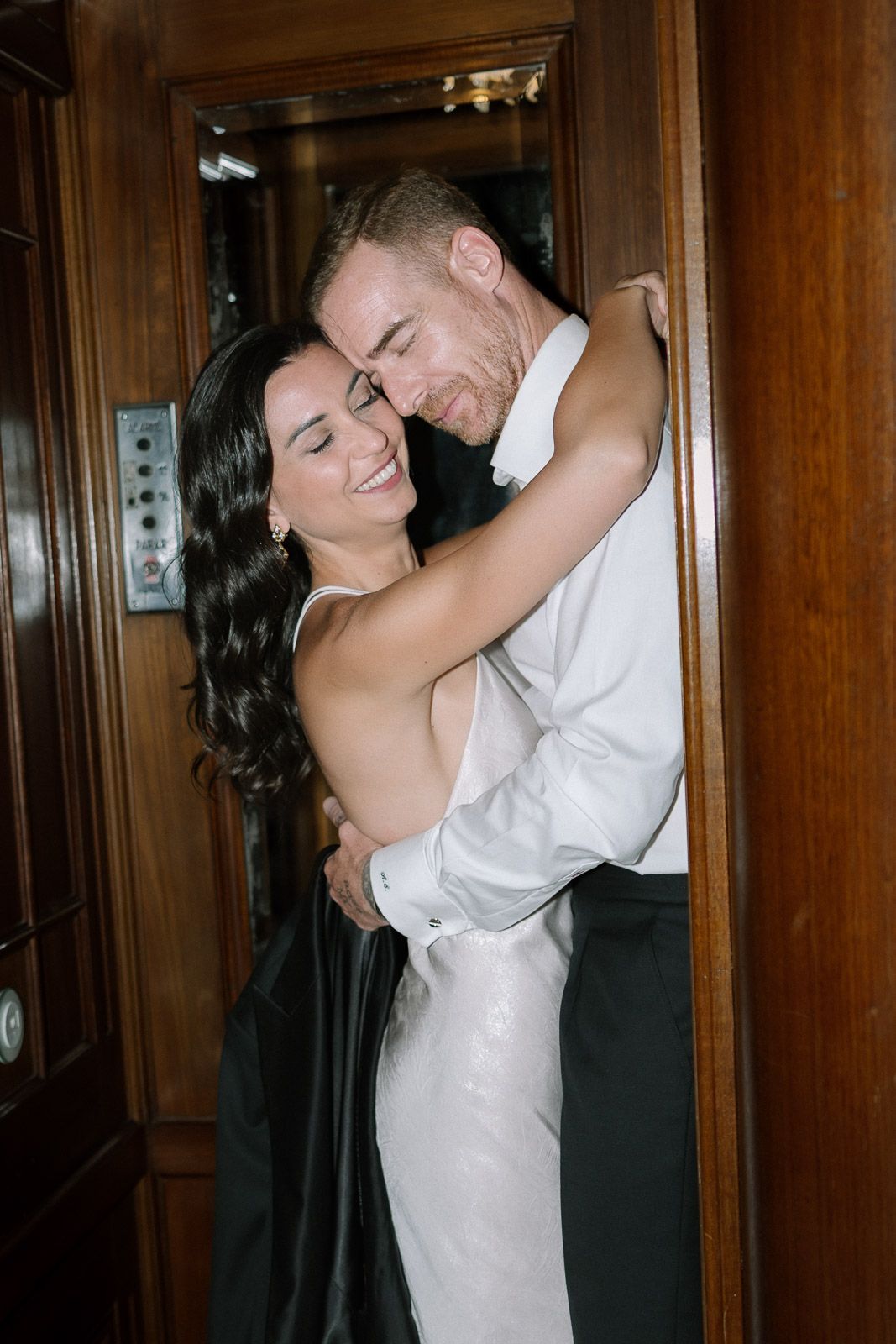 Bride and groom embracing and smiling in a wooden elevator doorway on their wedding day.