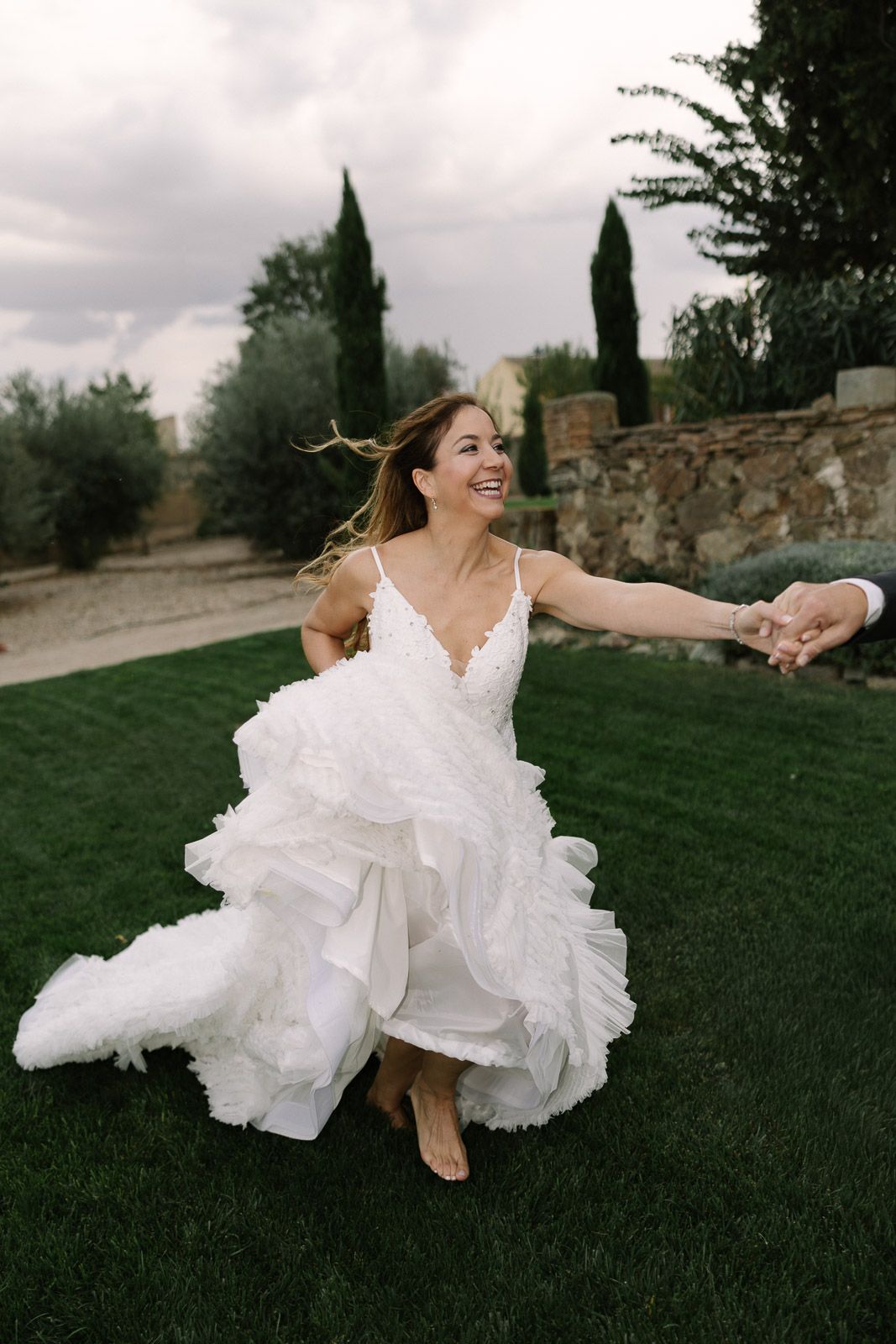 Joyful bride in a white ruffled wedding gown dancing barefoot on green grass, holding hands with groom outdoors.