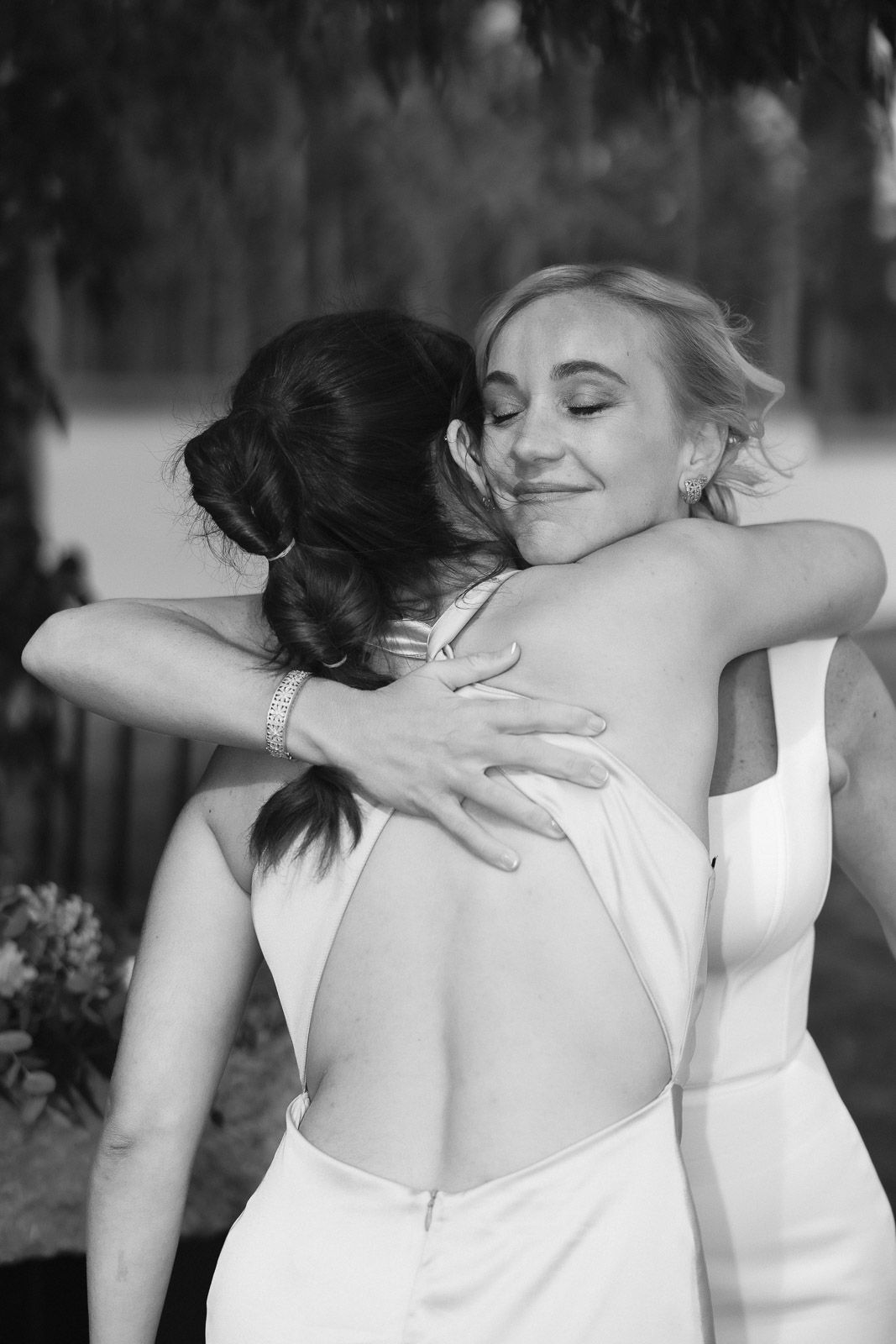 Two brides share an emotional hug in black and white wedding photo, one in open-back gown smiling warmly.