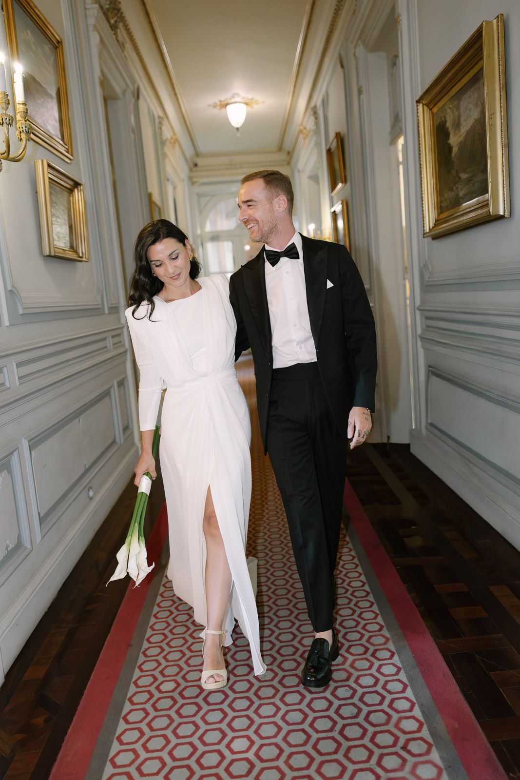 Elegant bride and groom walking together in ornate hallway, bride holding white calla lily bouquet.