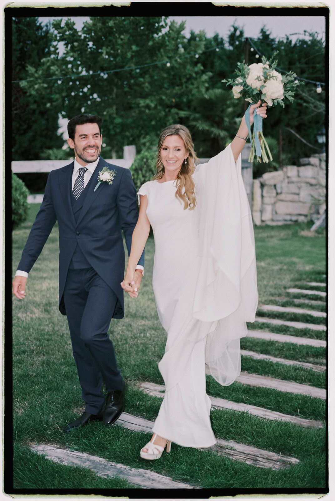 Happy bride and groom walking hand in hand outdoors, bride raising bouquet in white gown, groom in navy suit.