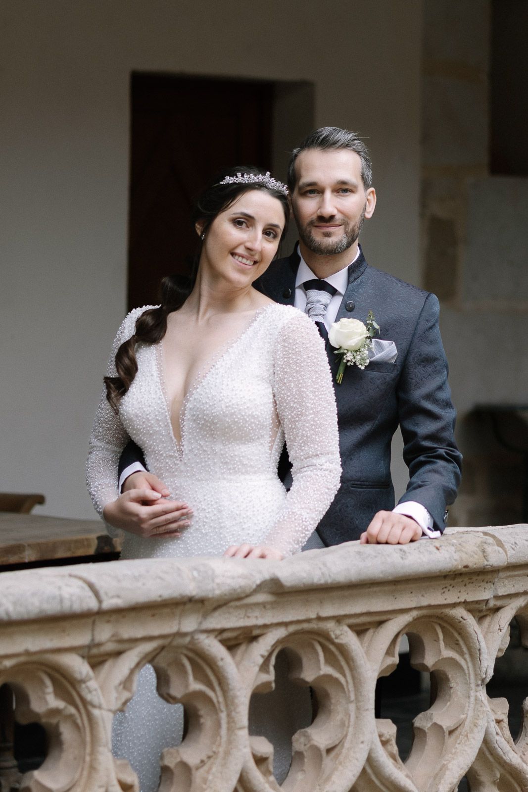 Elegant bride and groom posing on ornate stone balcony, bride in pearl gown with tiara, groom in gray suit.