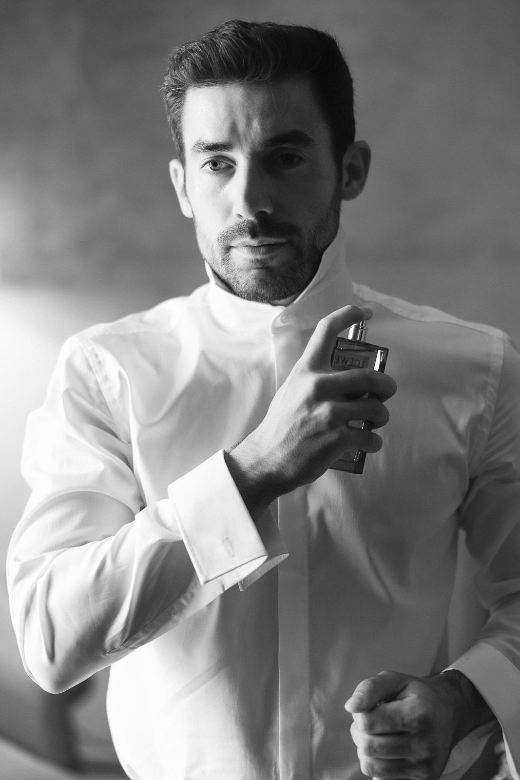 Black and white photo of a well-dressed man in white shirt applying cologne perfume bottle to his chest.