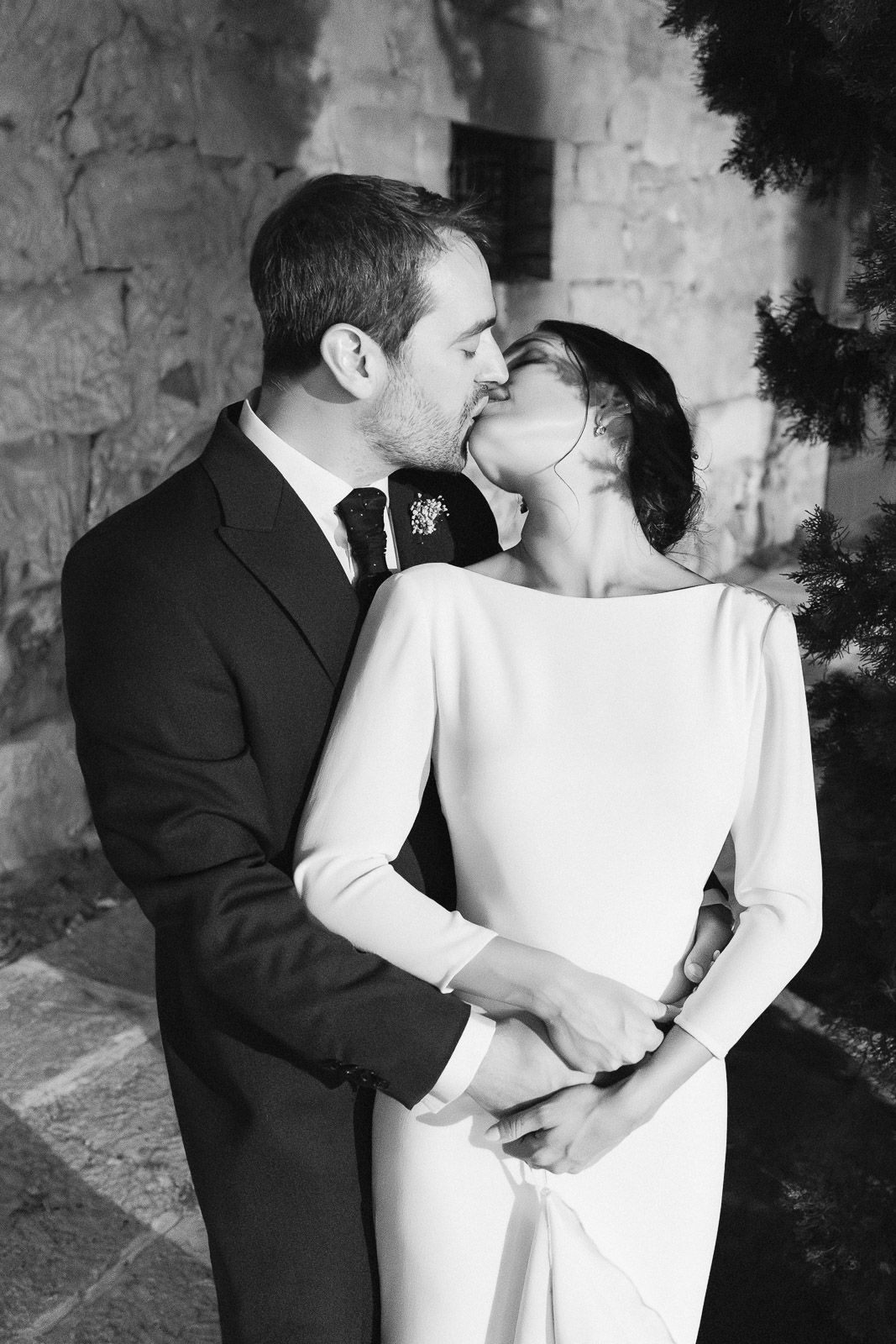 Black and white photo of a bride and groom sharing a romantic kiss on their wedding day near a stone wall.
