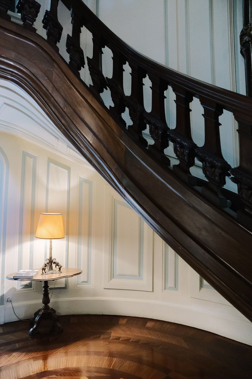 Elegant curved wooden staircase with ornate dark balustrade beside a vintage lamp on marble table in classic interior.
