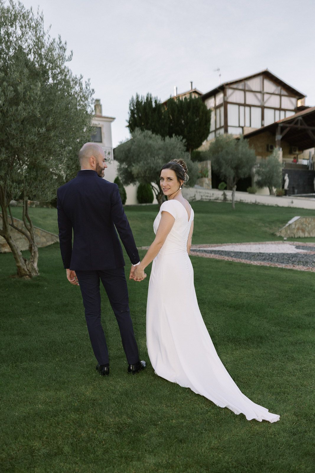 Bride in white gown holds groom's hand on green lawn, turning to smile, with rustic buildings in background.