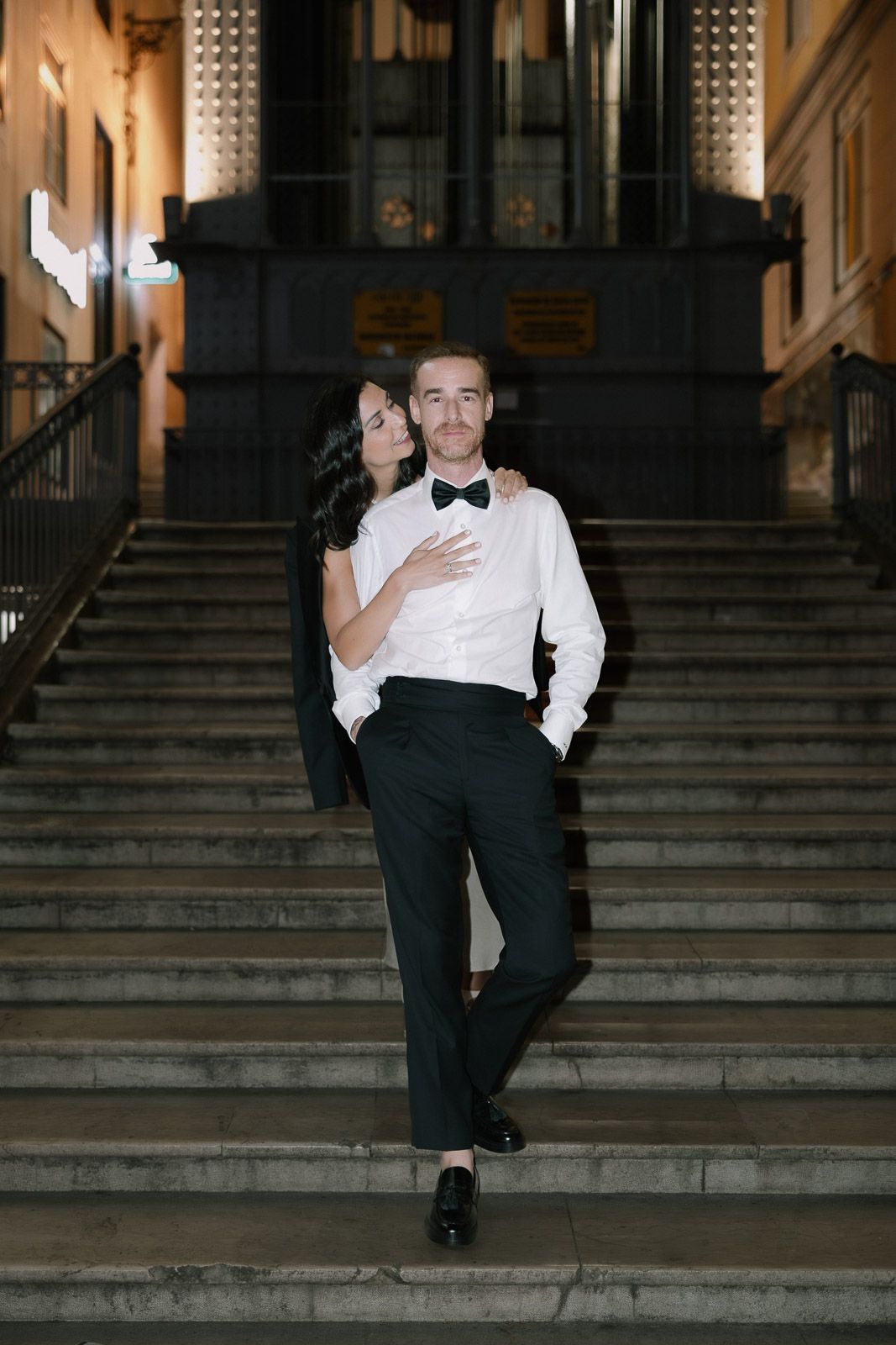 Couple in formal attire embracing on grand staircase at night, man in tuxedo with bow tie.