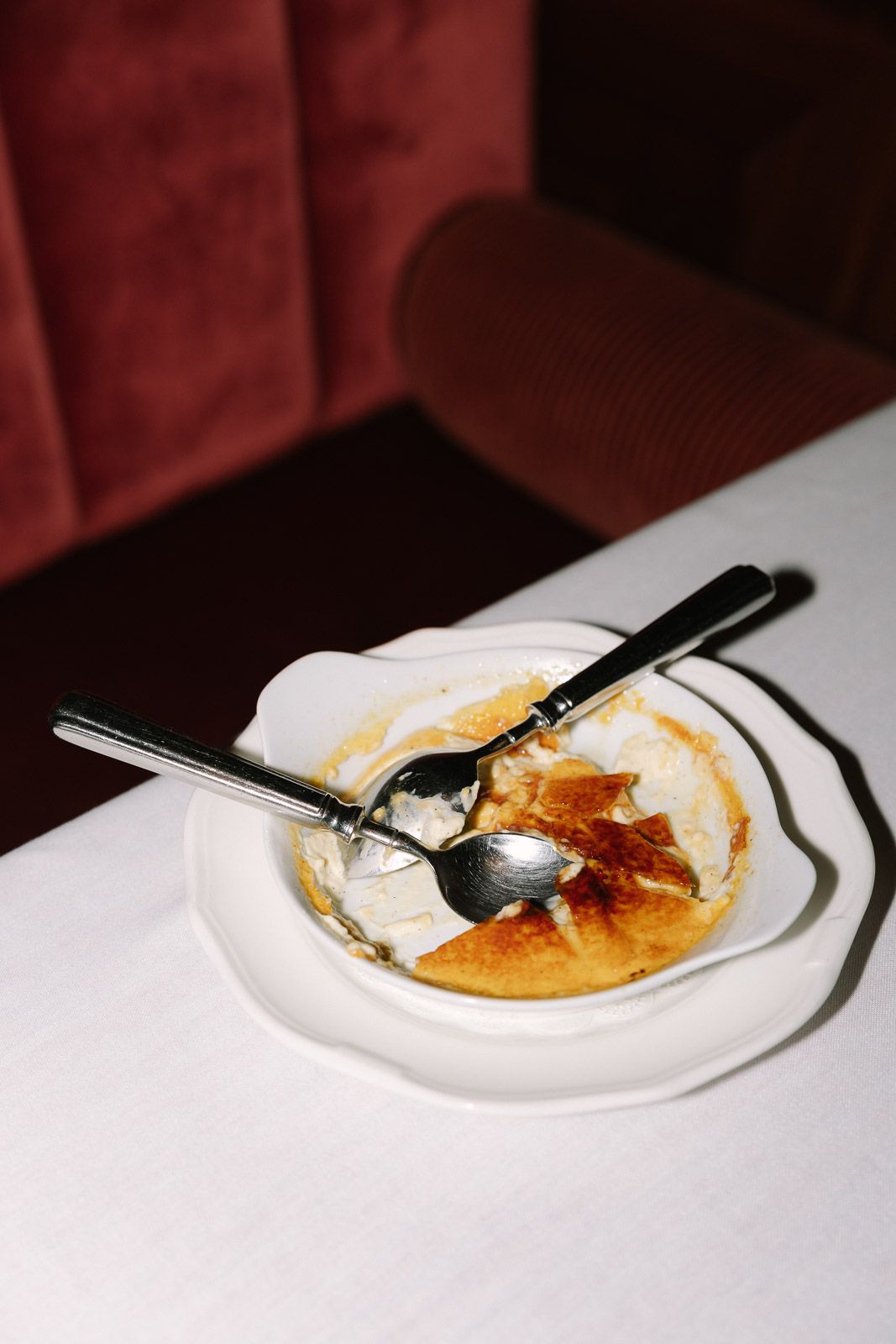 Nearly empty crème brûlée dessert dish with two spoons on white plate at restaurant table.