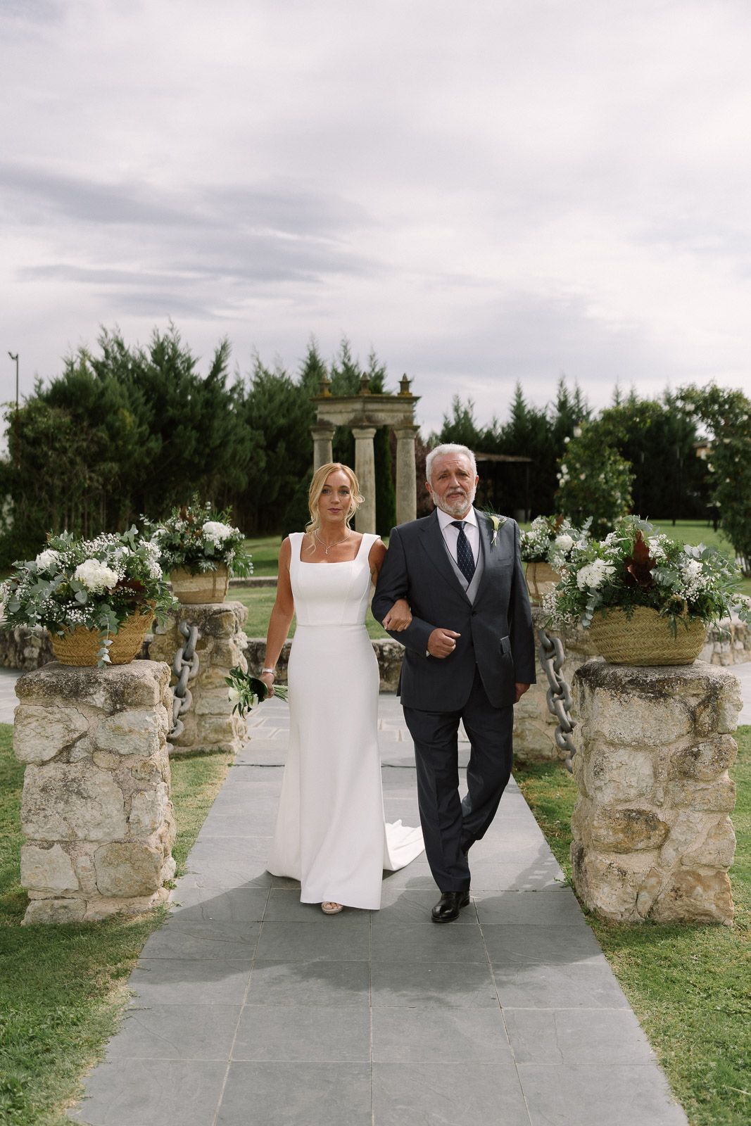 Bride in white gown walks down outdoor aisle with older man in dark suit at elegant garden wedding venue.