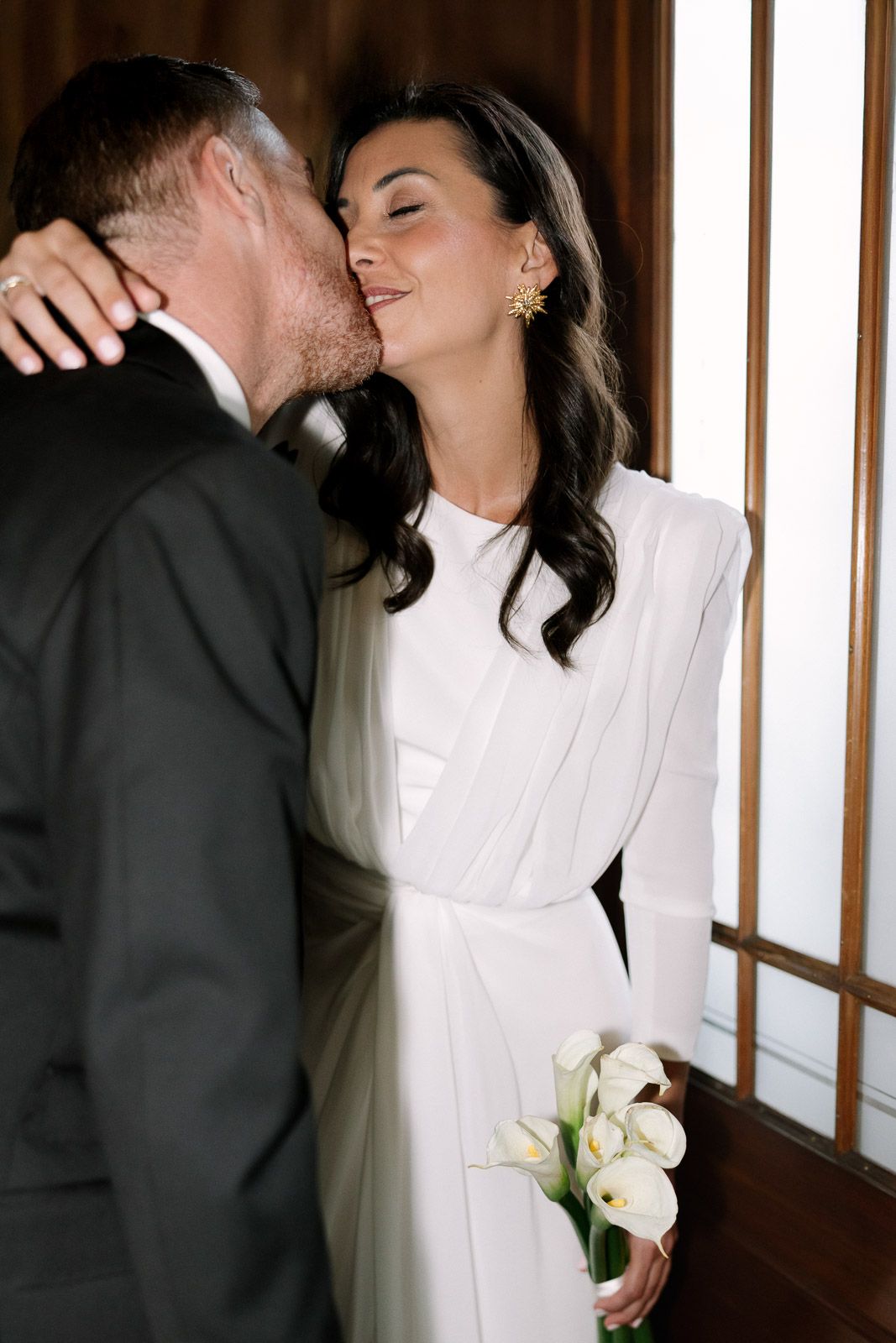 Bride in white long-sleeve gown holding calla lilies receives a kiss from groom in black suit near wooden doors.
