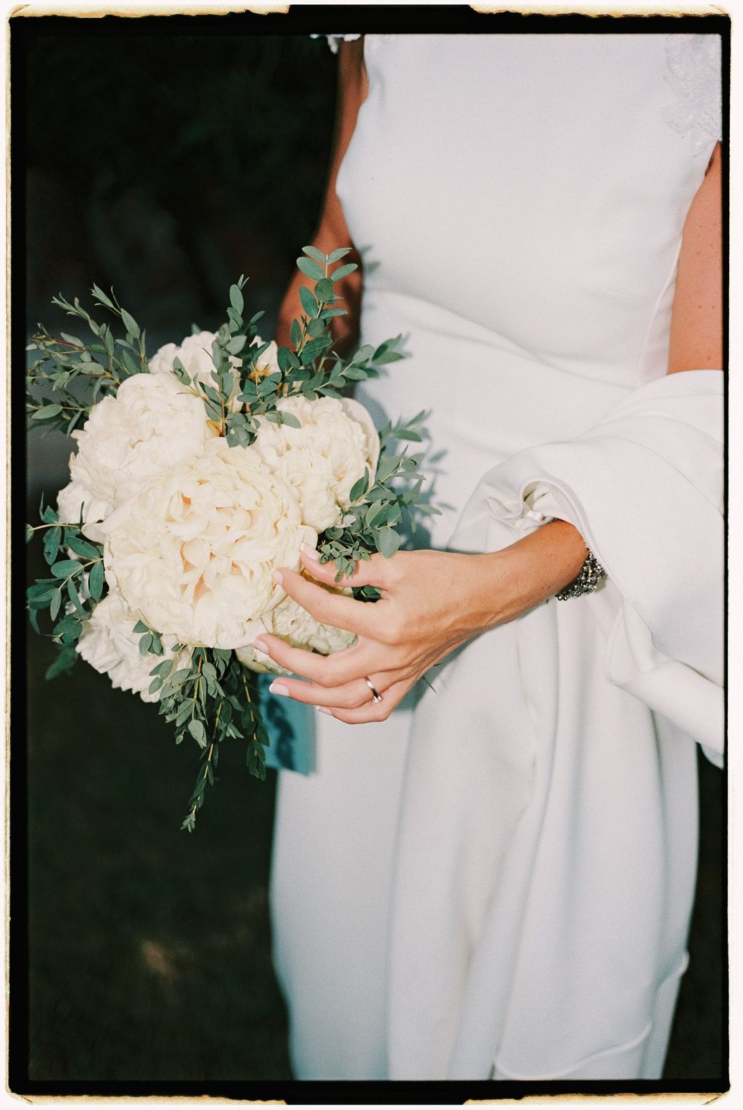 Bride holding a white peony and eucalyptus wedding bouquet in a white dress with long sleeves.