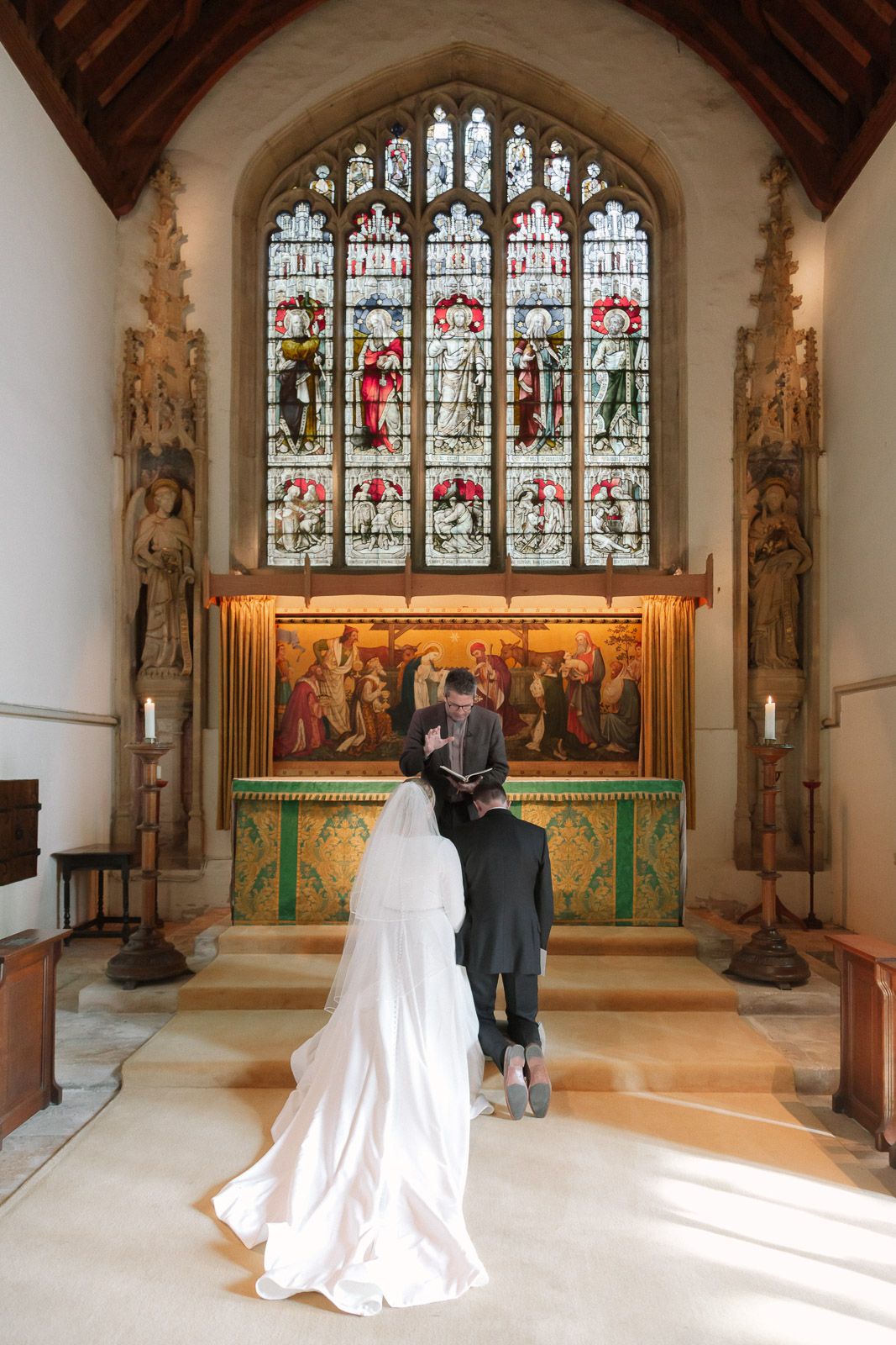 Bride and groom stand at church altar before stained glass window during traditional wedding ceremony.