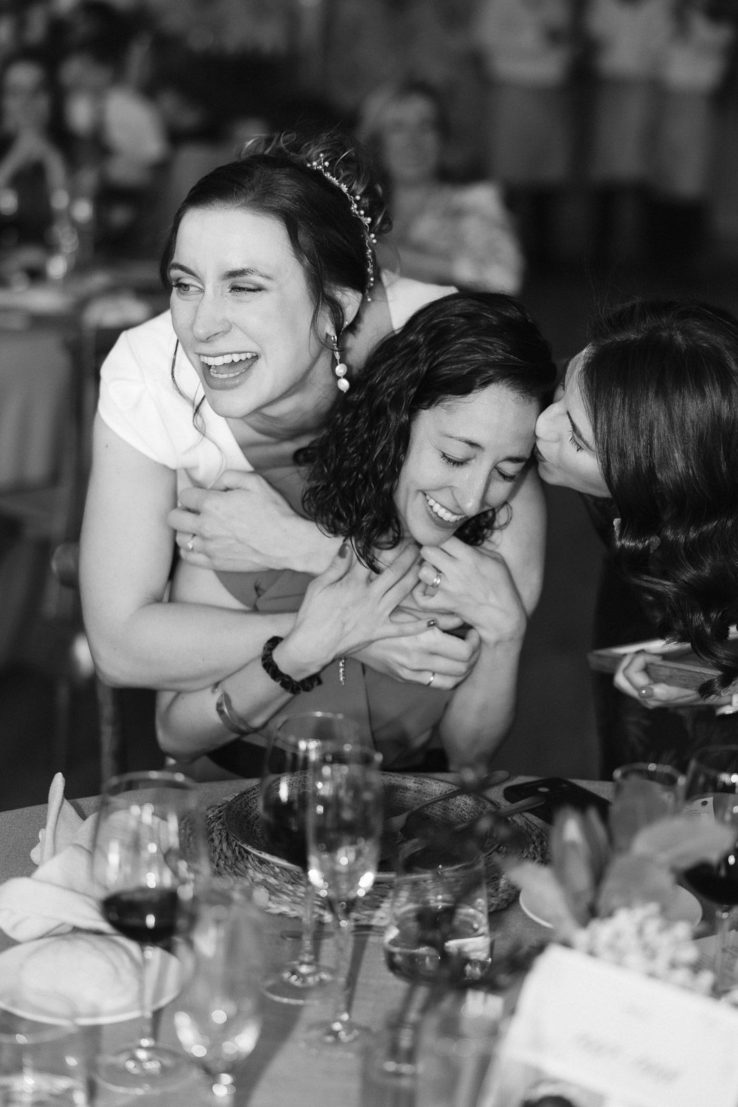 Black and white photo of three women laughing and embracing joyfully at a wedding reception dinner table.