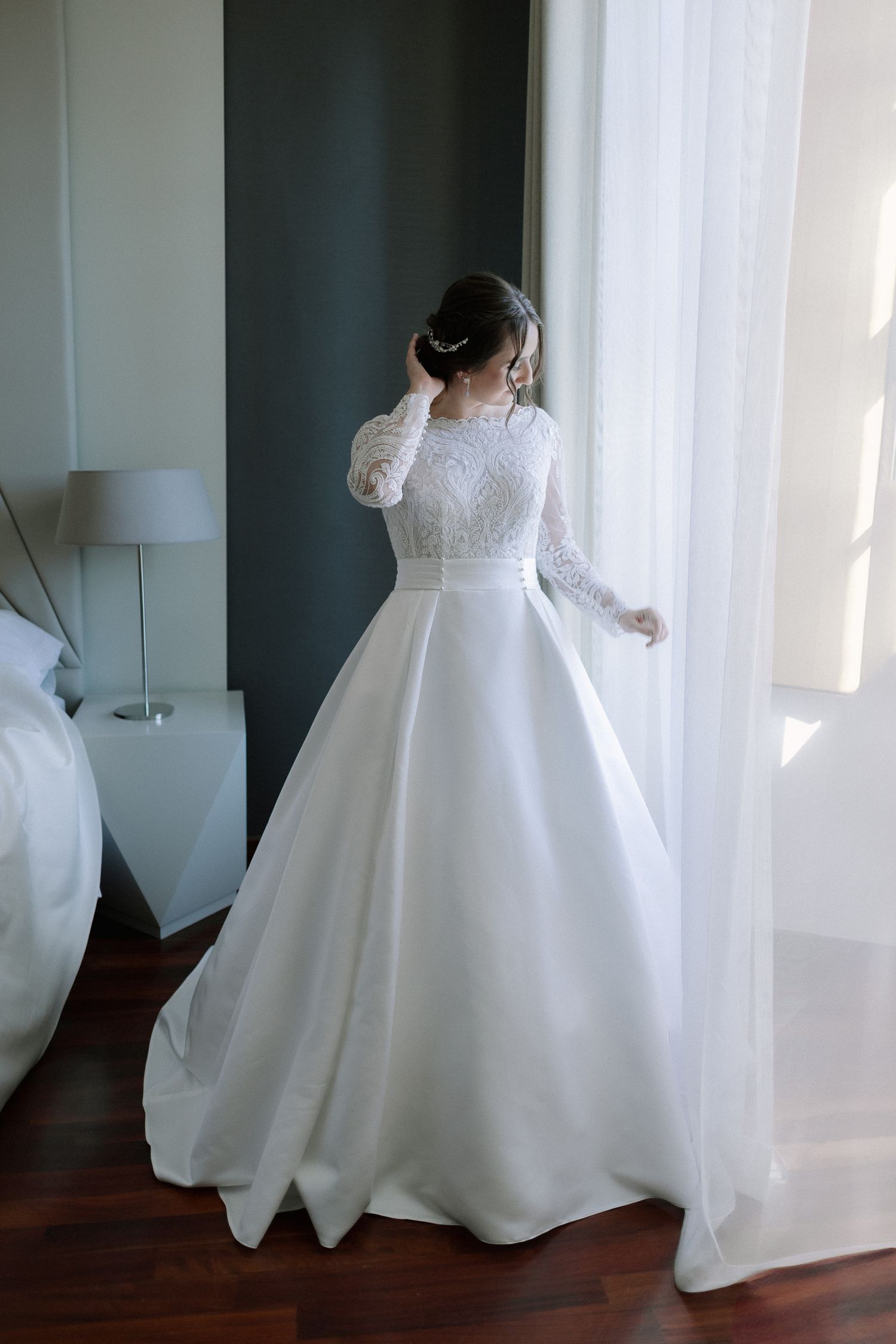 A bride in a flowing white wedding gown twirls gracefully on a hardwood floor in natural light from nearby windows.
