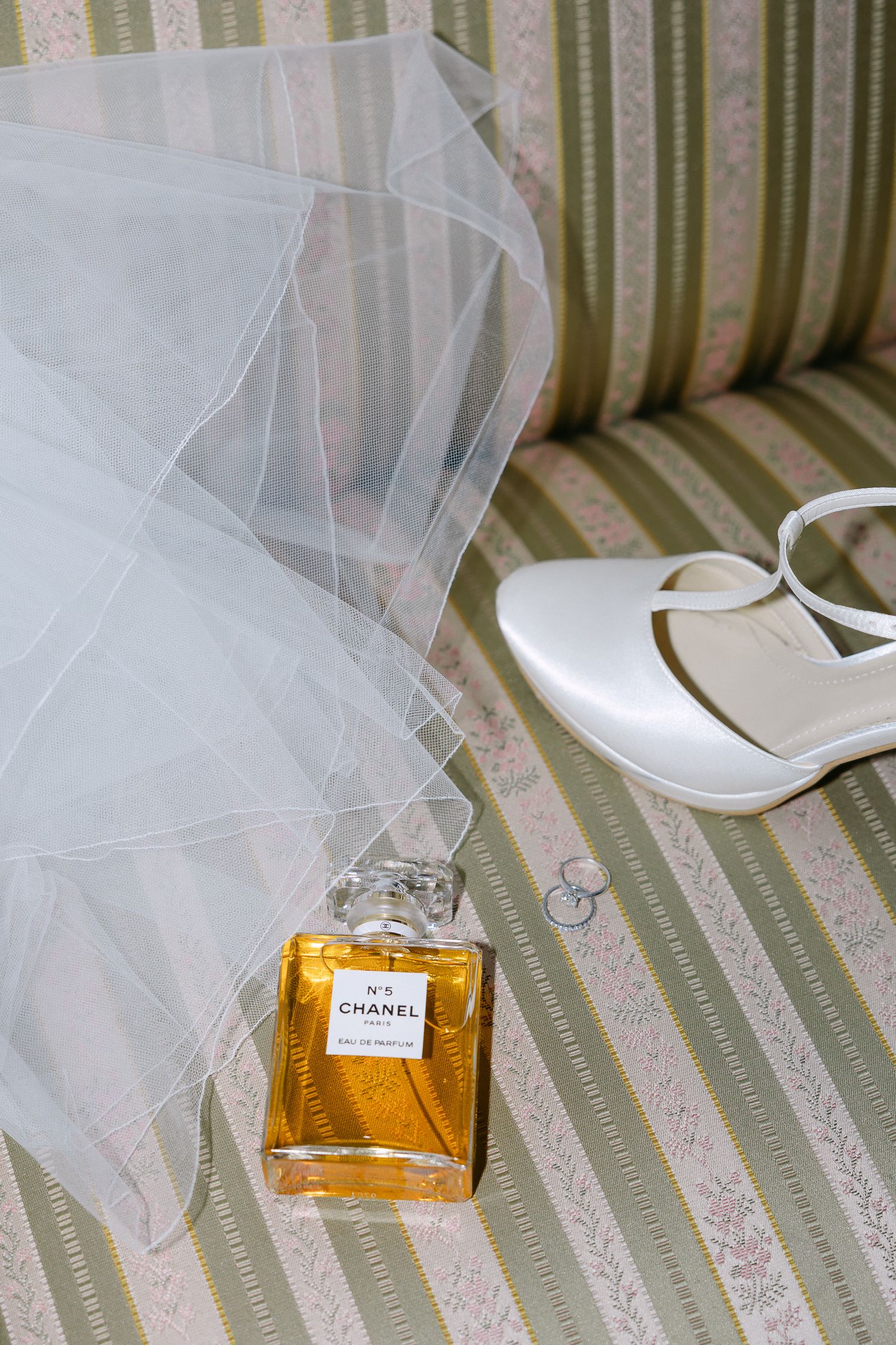 A bottle of Chanel perfume rests on a striped couch next to white bridal shoes and delicate wedding veil.