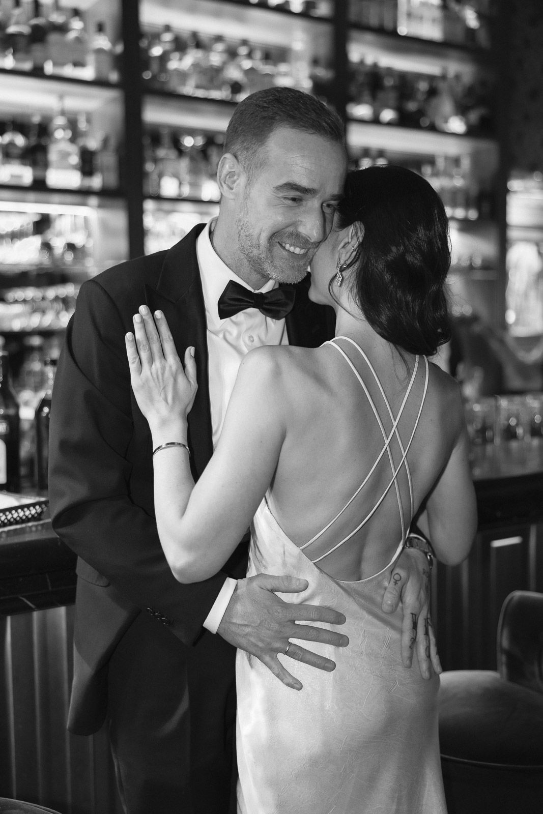Black and white photo of smiling groom in tuxedo embracing bride in backless wedding gown at bar.