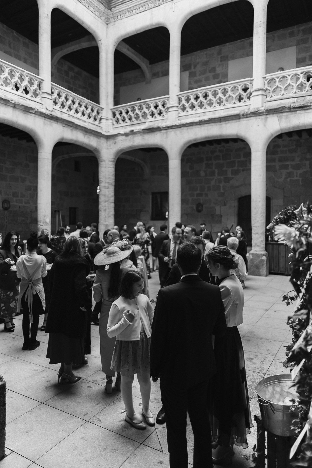 Black and white photo of elegantly dressed guests mingling in a historic stone courtyard with arched colonnades.