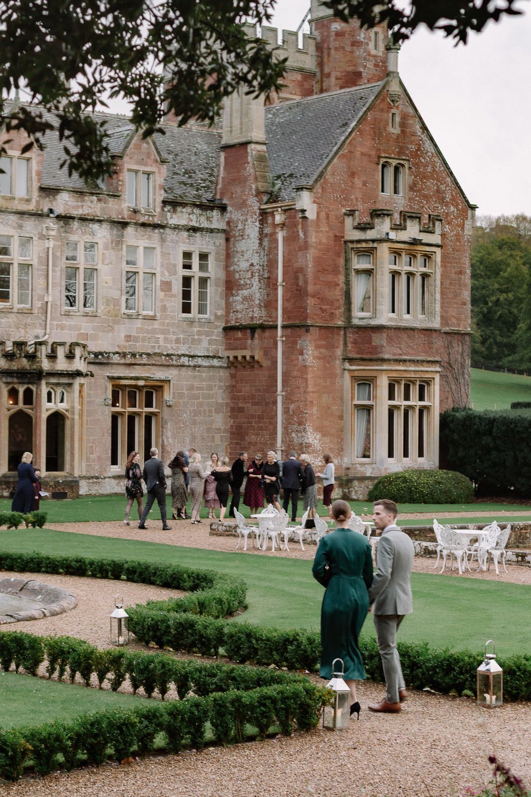 Elegant English manor house wedding venue with manicured gardens, lanterns, and guests mingling on the grounds.