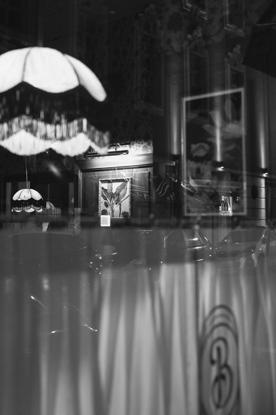 Black and white photo of a restaurant interior reflected in glass, featuring vintage lamps and framed artwork.