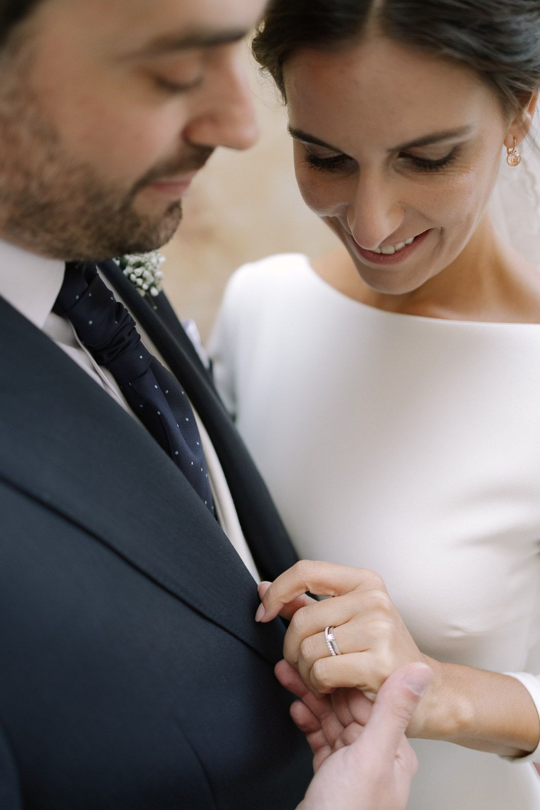 Bride in white gown smiles while adjusting groom's suit jacket, showcasing her wedding ring during ceremony.