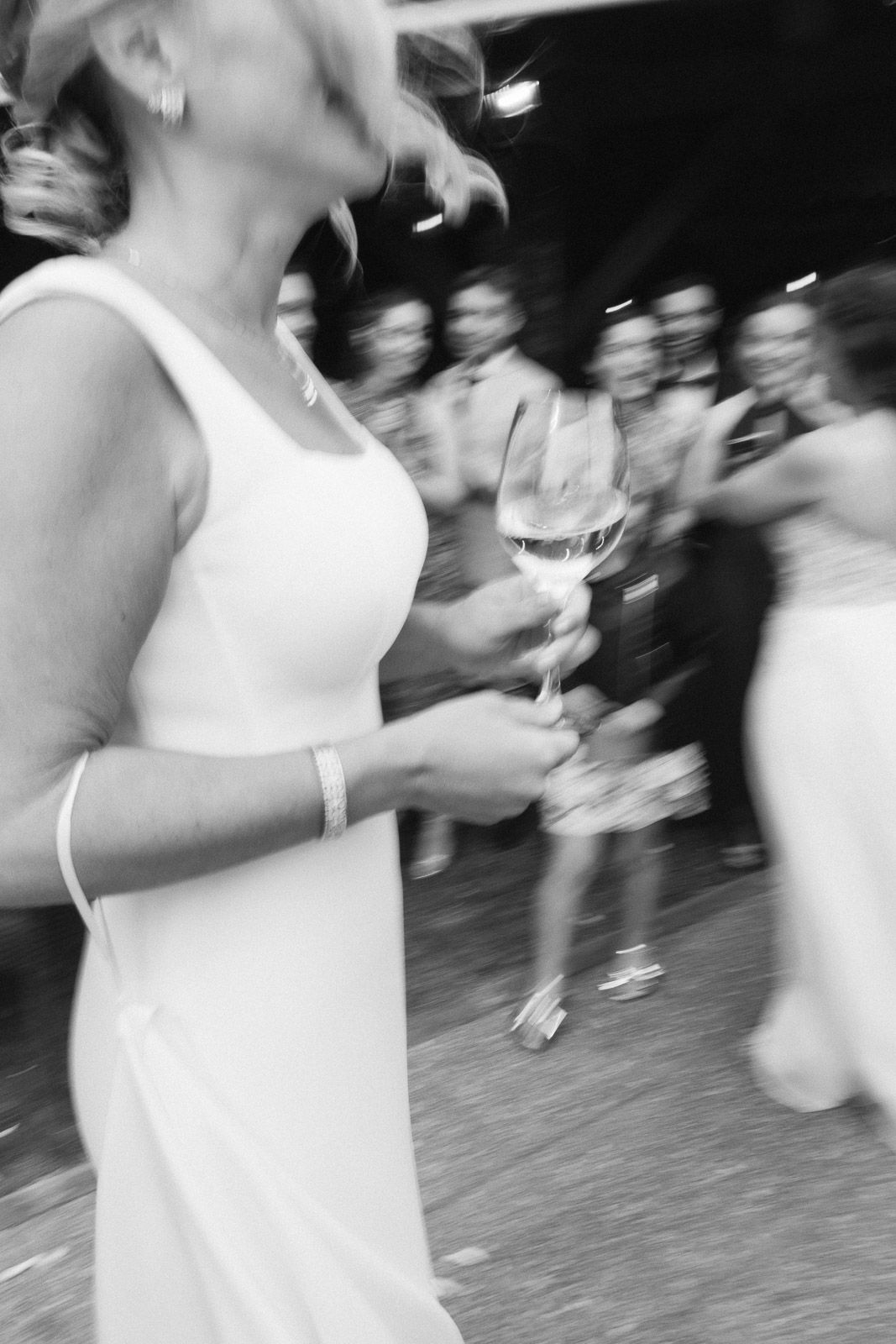 Blurred black and white photo of a bride in white dress holding a wine glass while dancing.