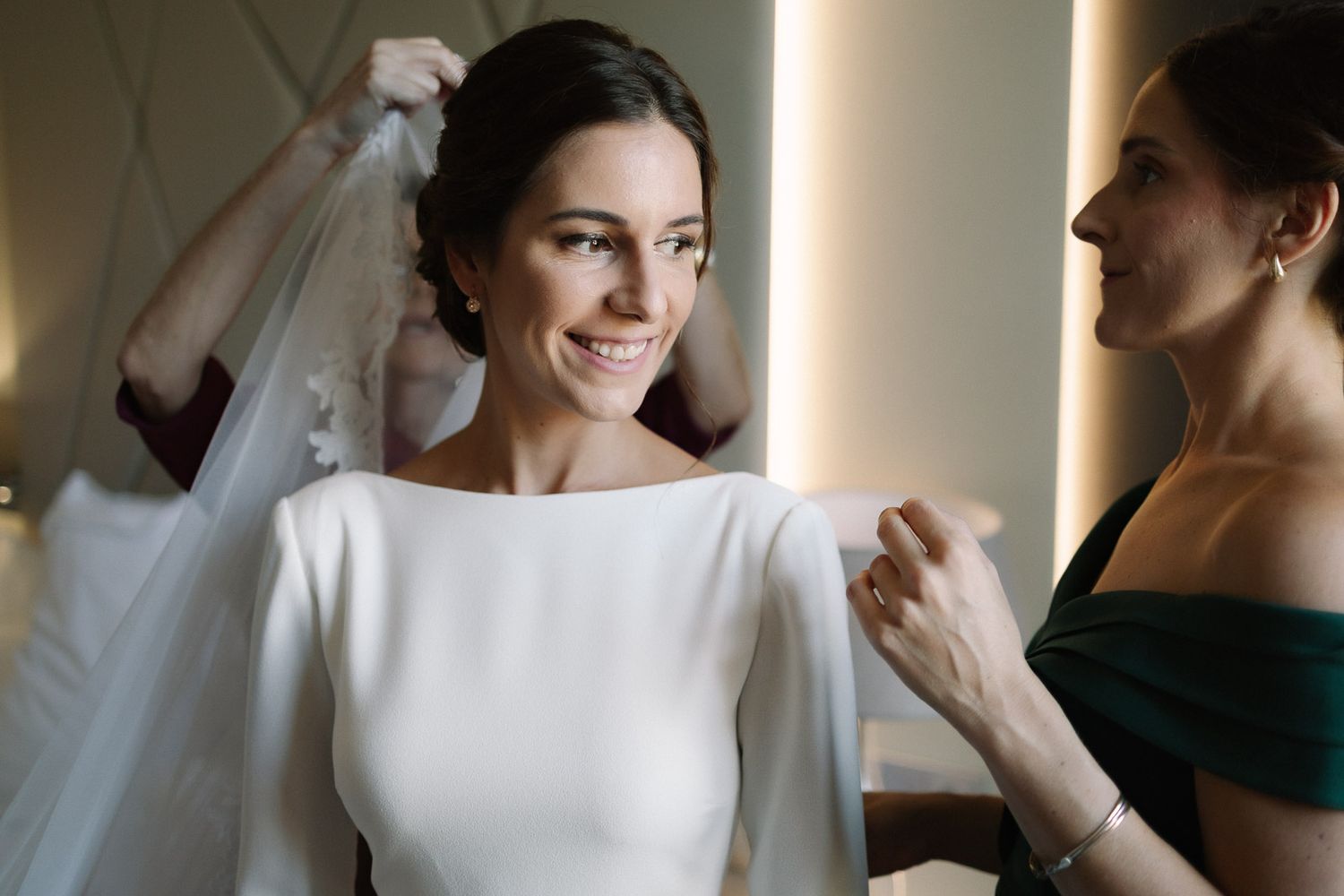 Bride smiling as bridesmaid helps adjust her white wedding veil and elegant long-sleeve bridal gown.