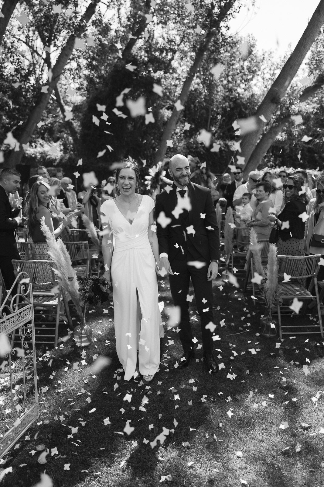 Black and white photo of a joyful bride and groom walking down the aisle as confetti fills the air at an outdoor wedding.