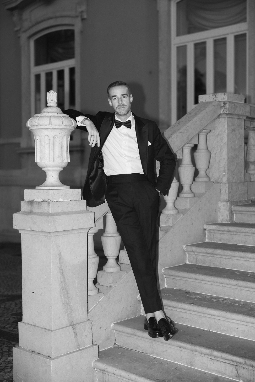 Elegant man in tuxedo posing on marble staircase in black and white photo.