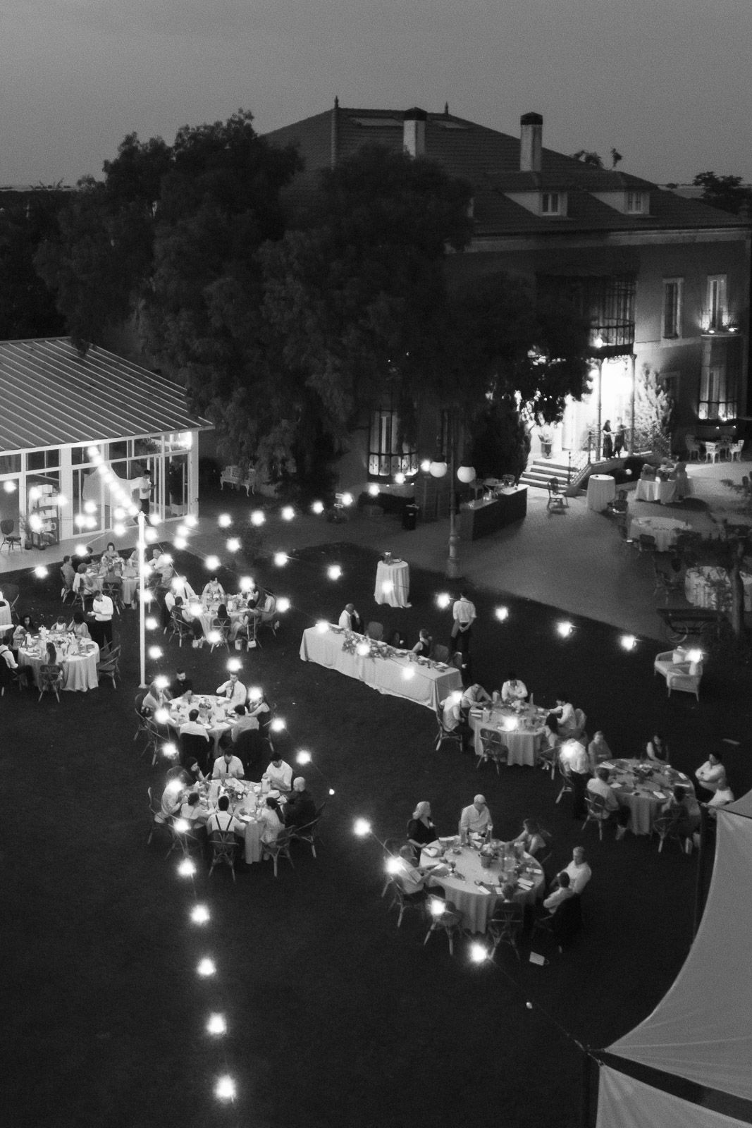 Aerial black and white photo of an elegant outdoor evening event with string lights and round dining tables.